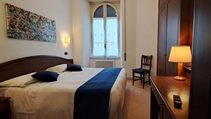 A hotel room with a bed, a chair, a desk with a lamp, a window, and a painting on the wall. - Hotel Bonazzi (Perugia)