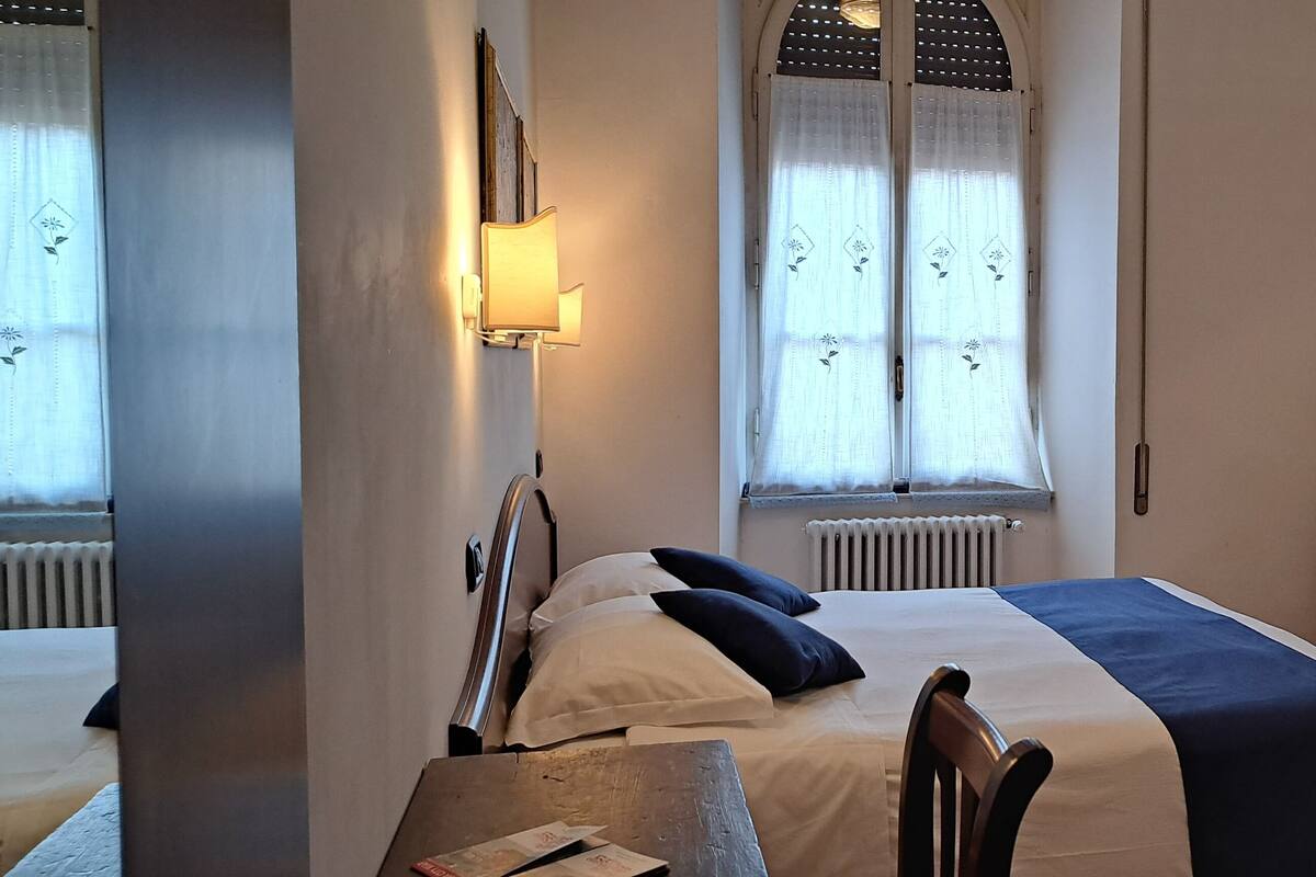 Habitación de hotel con cama, escritorio, silla, televisor, armario y ventana con cortinas.