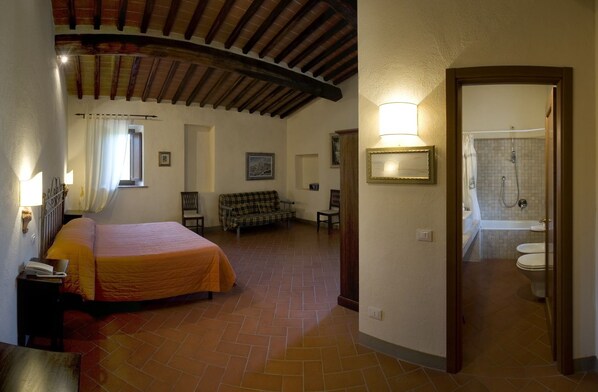 A bedroom with a bed, a nightstand, a lamp, a window with curtains, a sofa, and a bathroom with a bathtub and toilet. - Le Noci (Gaiole in Chianti)