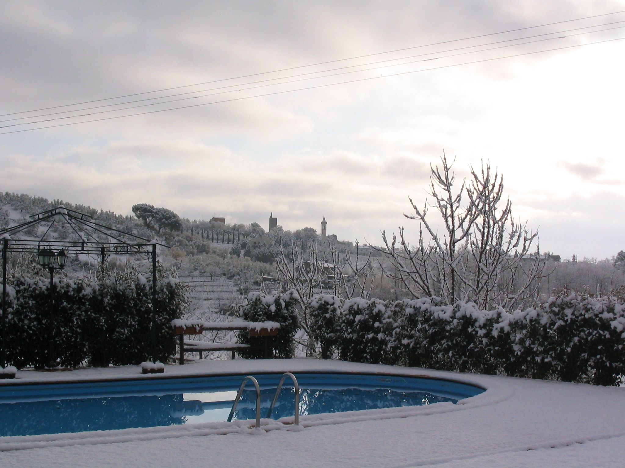 A snow-covered outdoor area with a swimming pool, a pergola, and a view of a distant town.