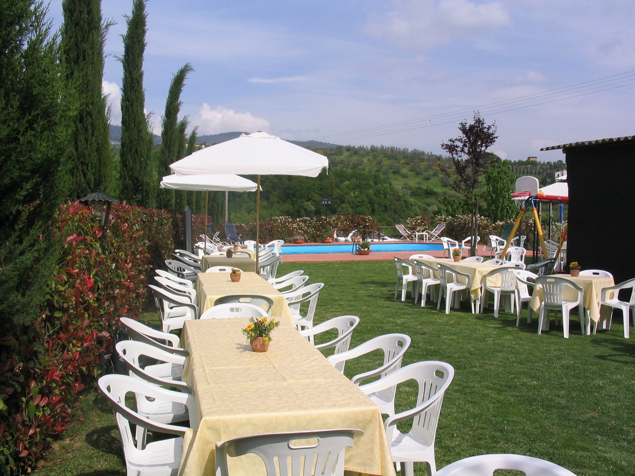 An outdoor dining area with white plastic chairs and tables, a pool, and a scenic hillside view.