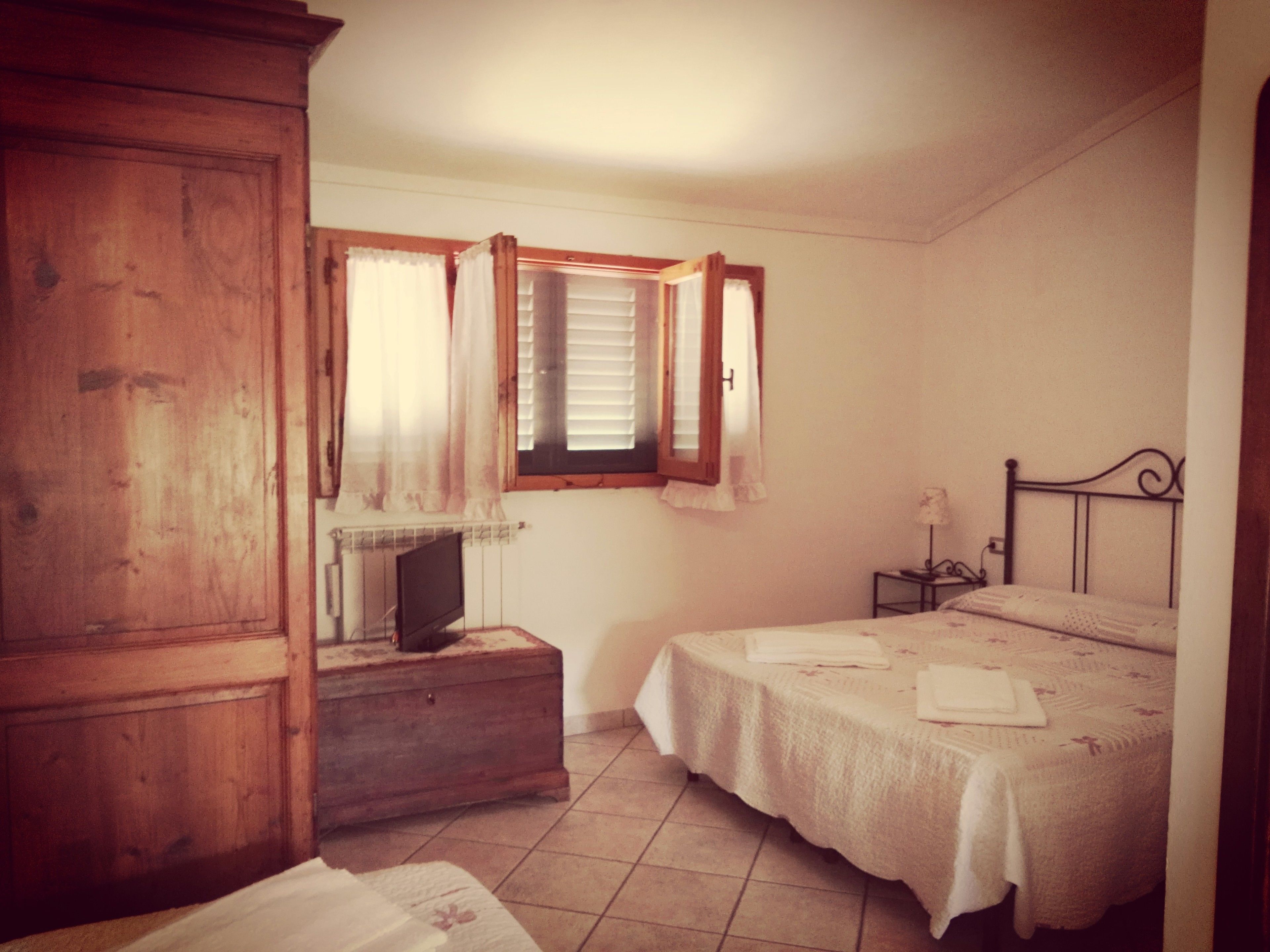 Un dormitorio con un armario de madera, una mesita de noche con lámpara, una cama con cubrecama blanco y una ventana con contraventanas abiertas.