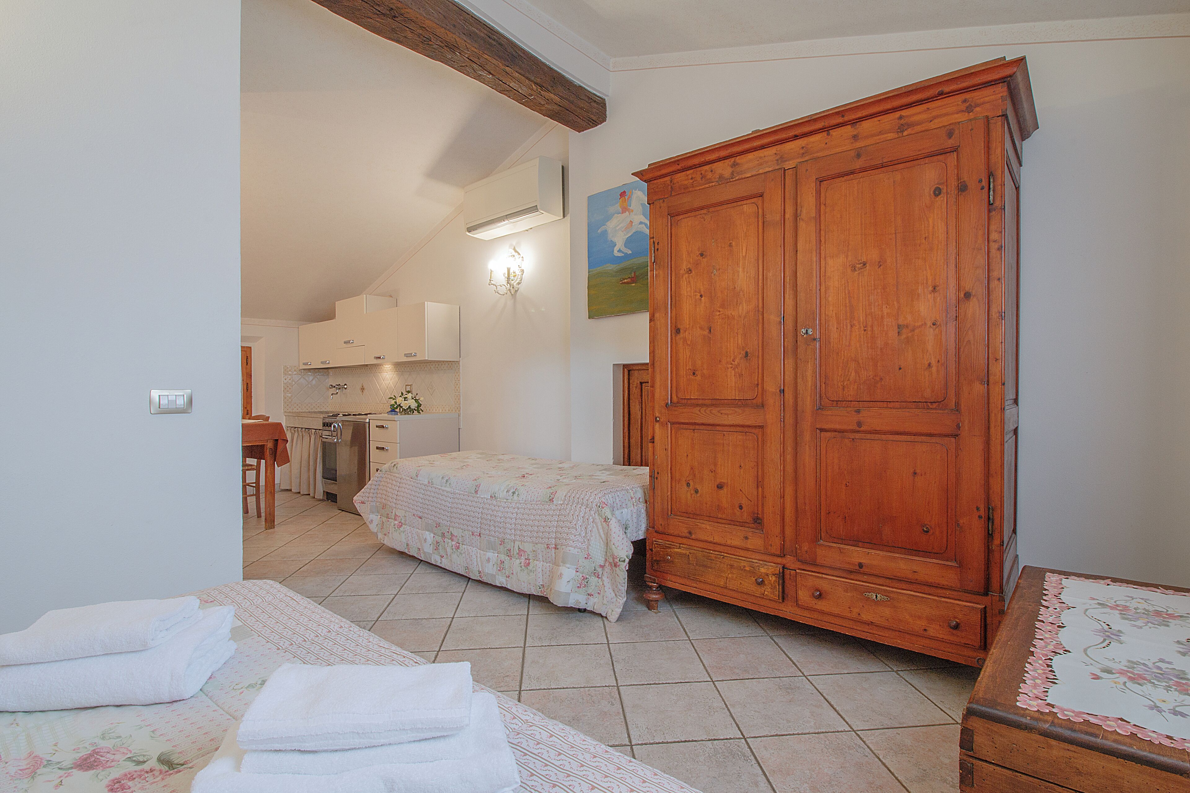 A bedroom with a wooden wardrobe, a bed with a floral bedspread, and a bathroom visible in the background.