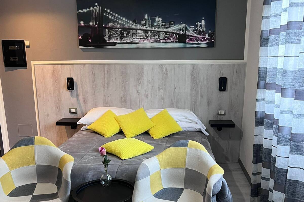 Una habitación de hotel moderna con una cama grande, dos sillas, una mesita con un florero y un cuadro de un paisaje urbano en la pared.
