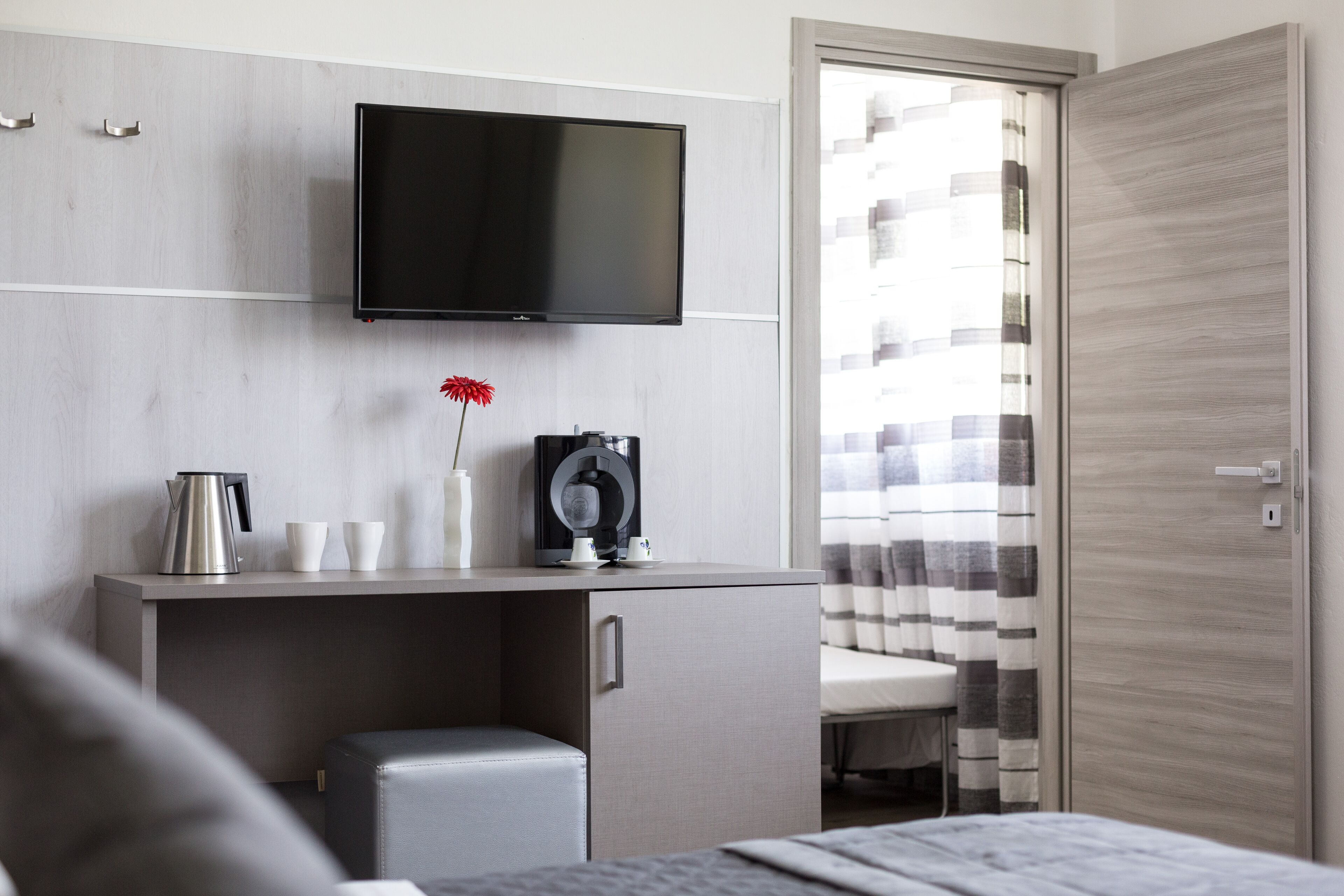 A modern hotel room with a flat-screen TV mounted on the wall, a small desk with a kettle, cups, and a camera, a cushioned bench, and a view of the outside through a window with striped curtains.