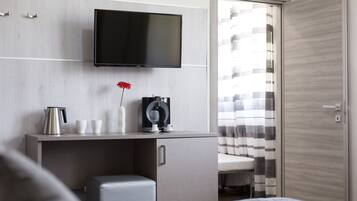 A modern hotel room with a flat-screen TV mounted on the wall, a small desk with a kettle, cups, and a camera, a cushioned bench, and a view of the outside through a window with striped curtains.