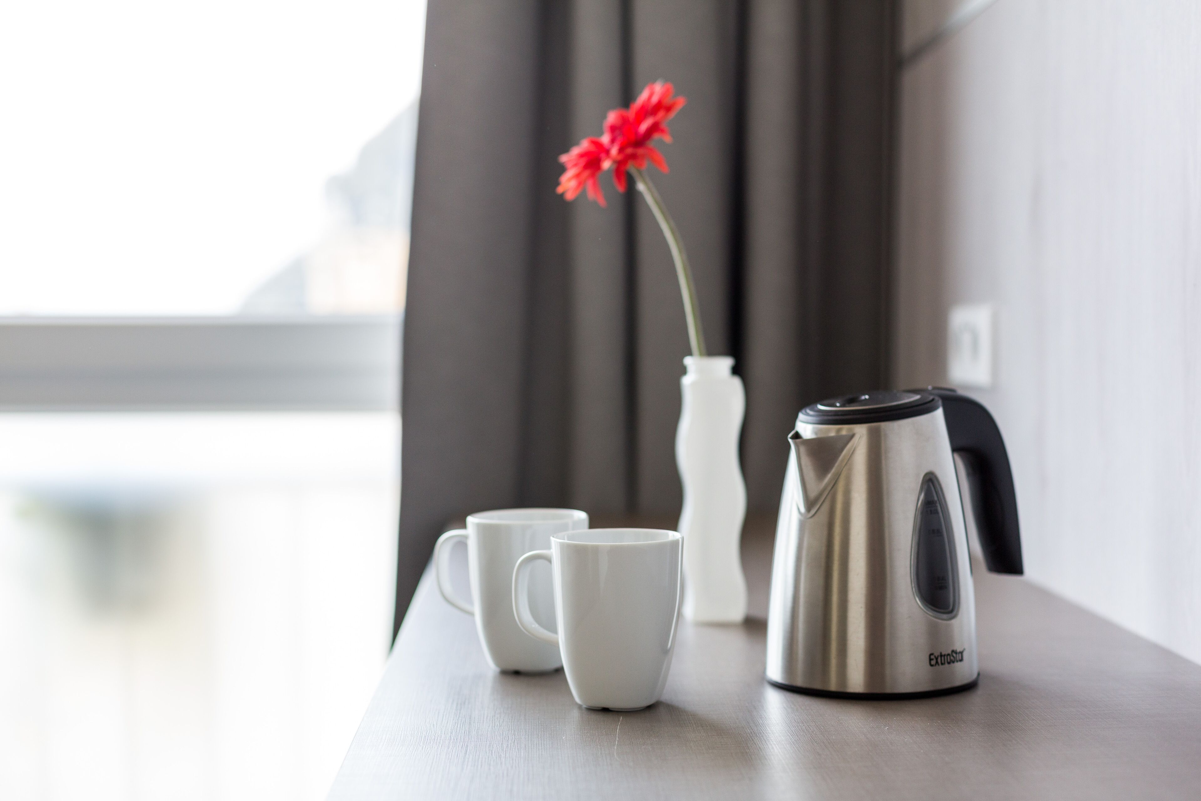 A table with a white vase containing a red flower, two white cups, and a stainless steel kettle.