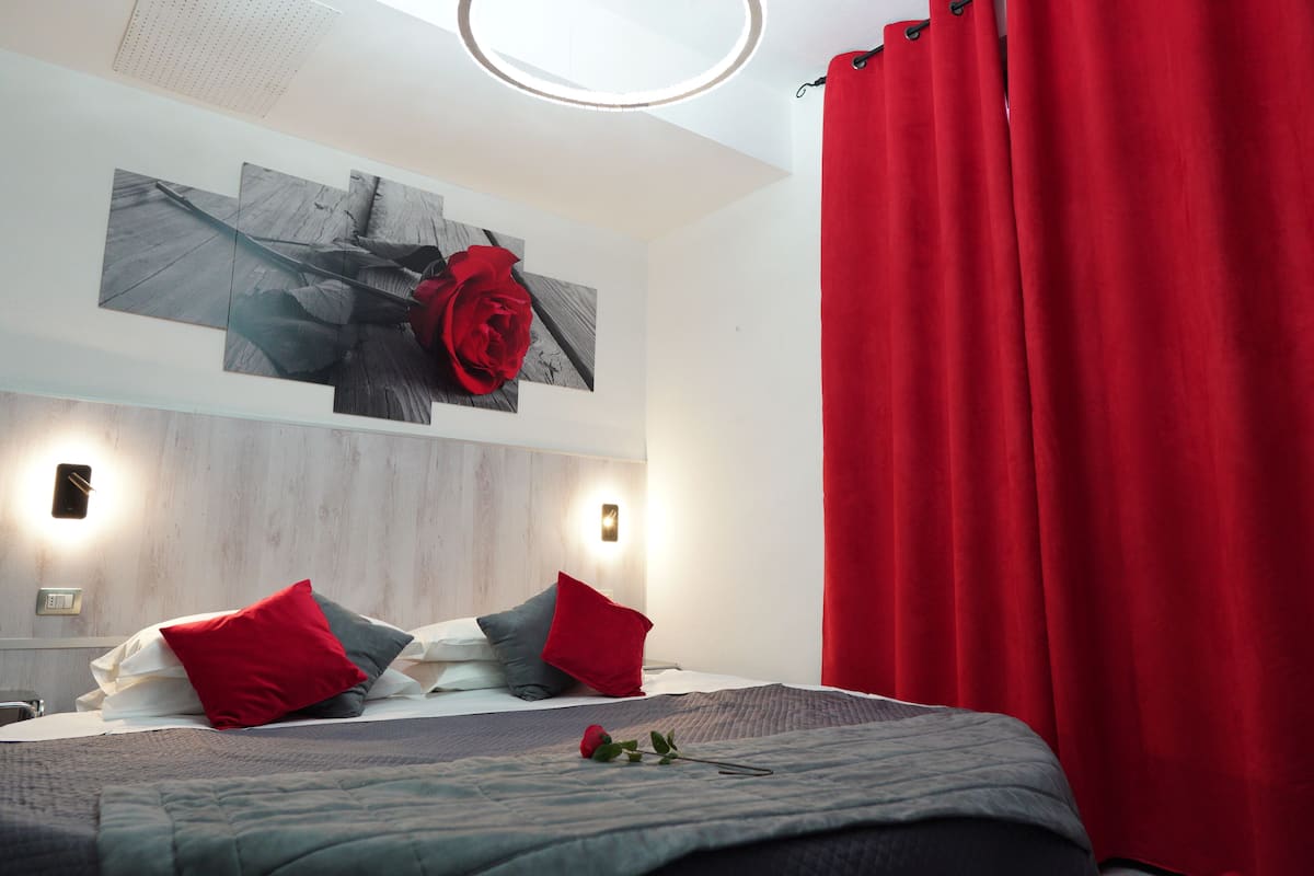 Una habitación de hotel moderna con una cama grande, almohadas rojas y grises, una cortina roja y un aplique de pared.