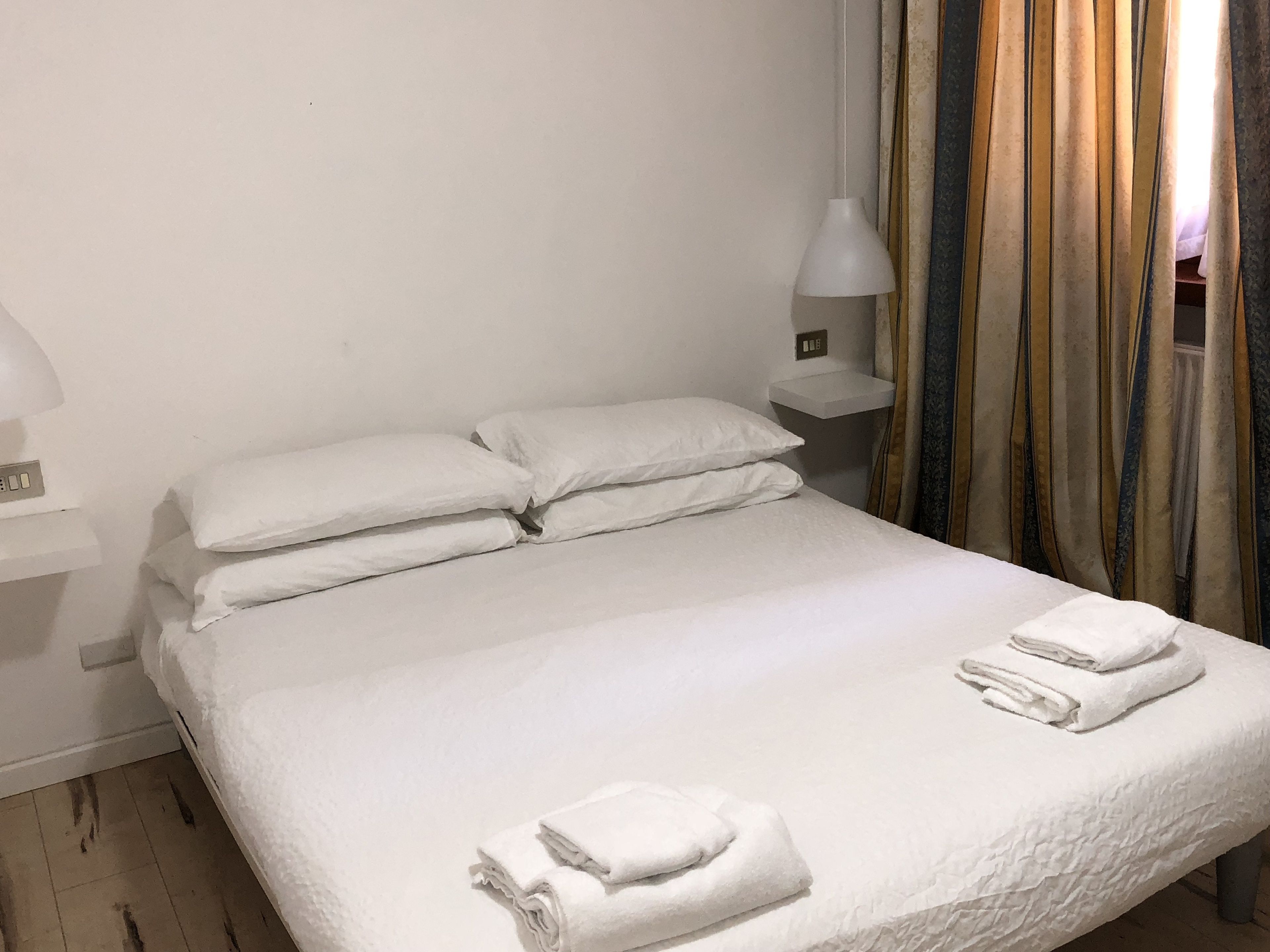 Una cama bien hecha con almohadas y toallas dobladas, una mesita de noche con lámpara y una cortina de estampado a rayas.