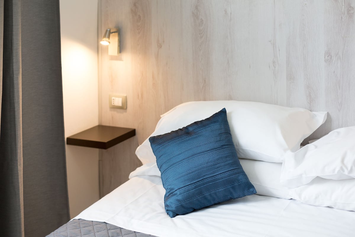 Una cama bien hecha con sábanas blancas y una almohada azul, una mesita de noche con lámpara y un panel de madera.
