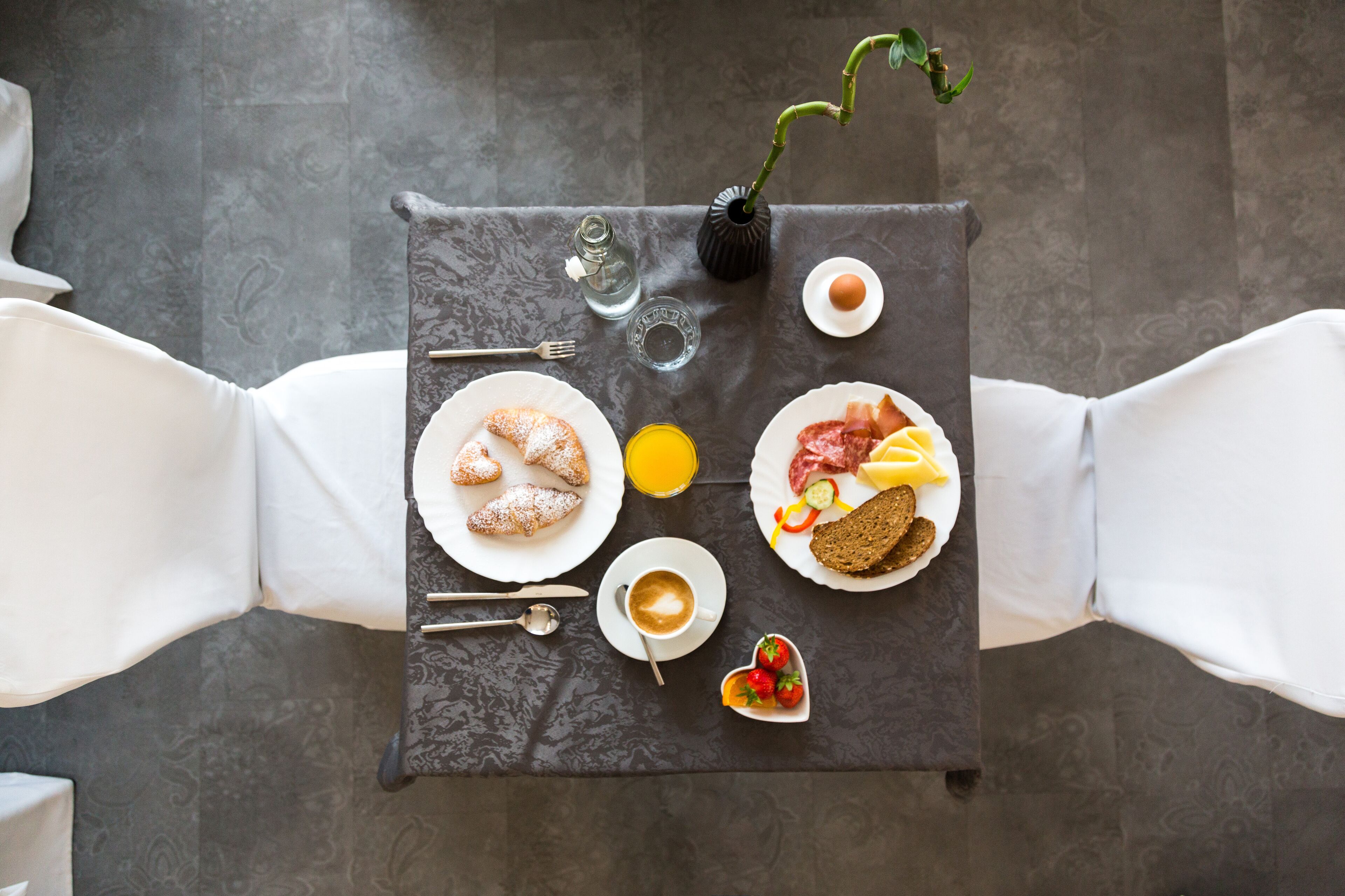 Una mesa de desayuno con cruasanes, huevos, tocino, tomates y un florero con plantas.