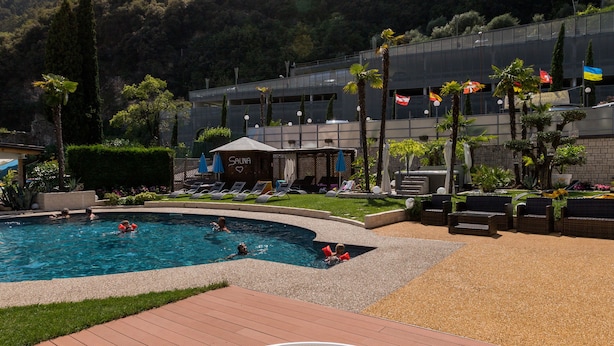 A hot tub on a wooden deck with a pool and lounge chairs, surrounded by greenery and mountains in the background.