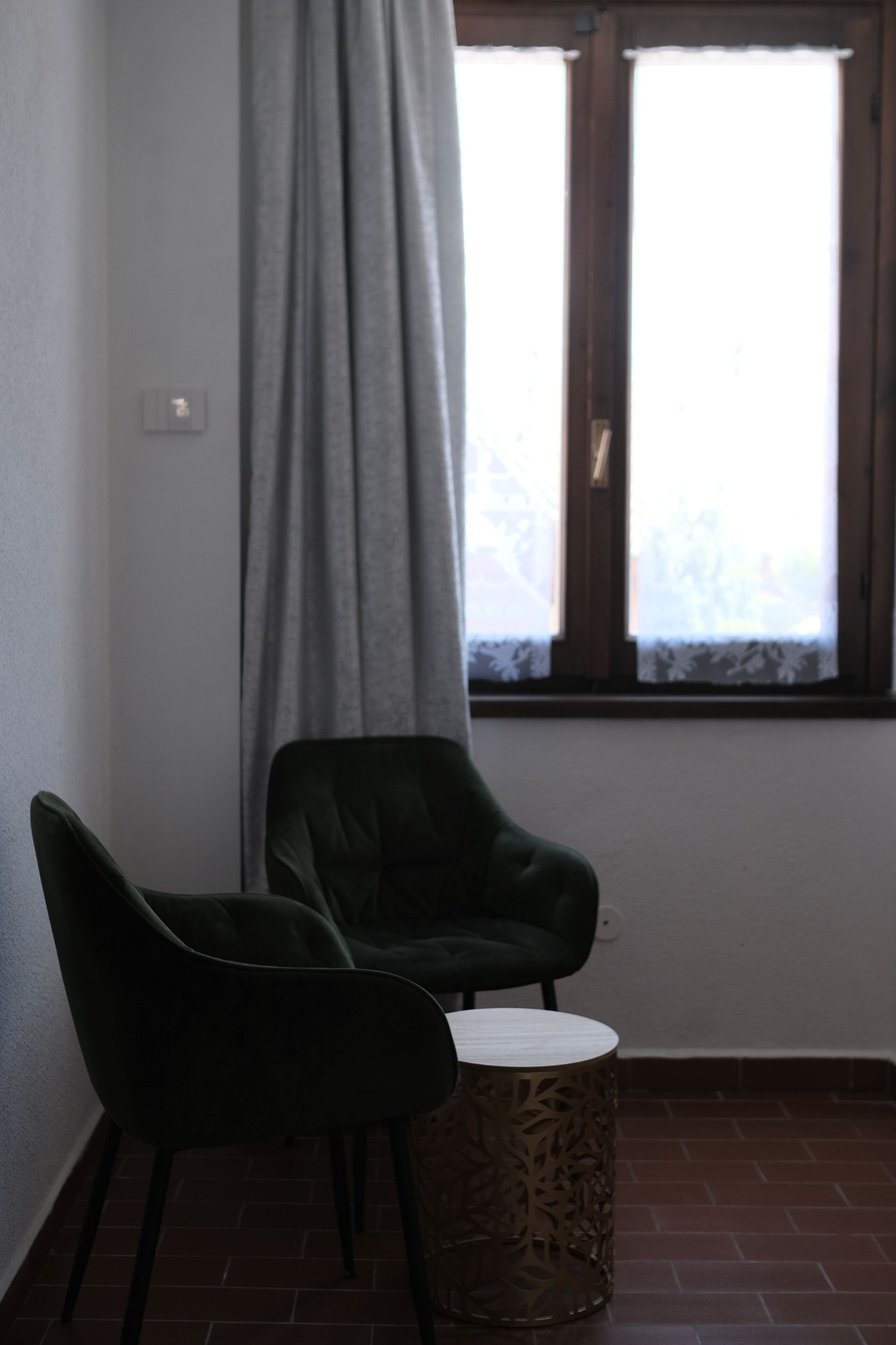 Une pièce avec un sol en briques, deux fauteuils vert foncé, une petite table ronde et une fenêtre avec des rideaux.