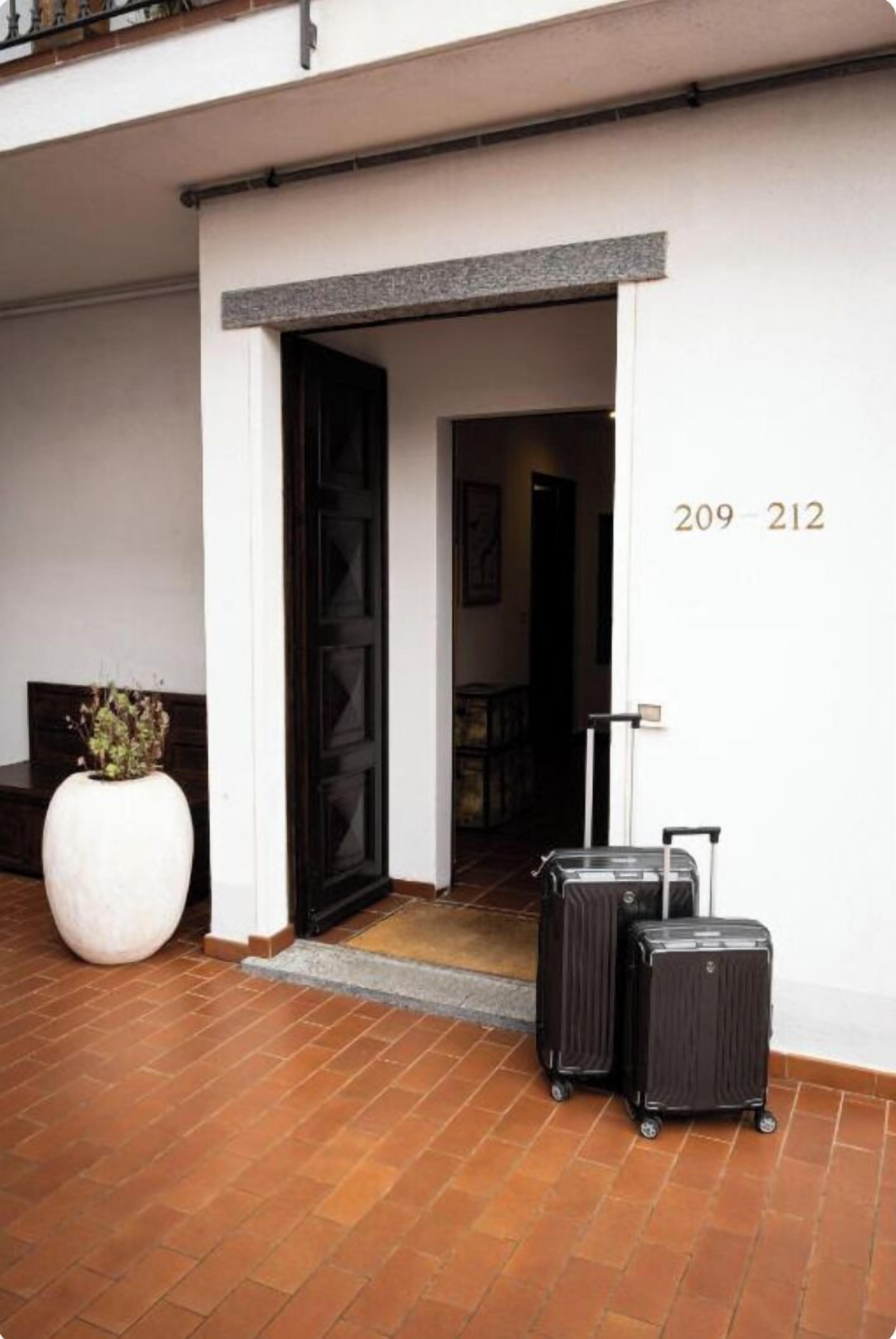 A hotel entrance with a white wall, a black door, and a number '209-212' on the wall. There are two suitcases in front of the door.