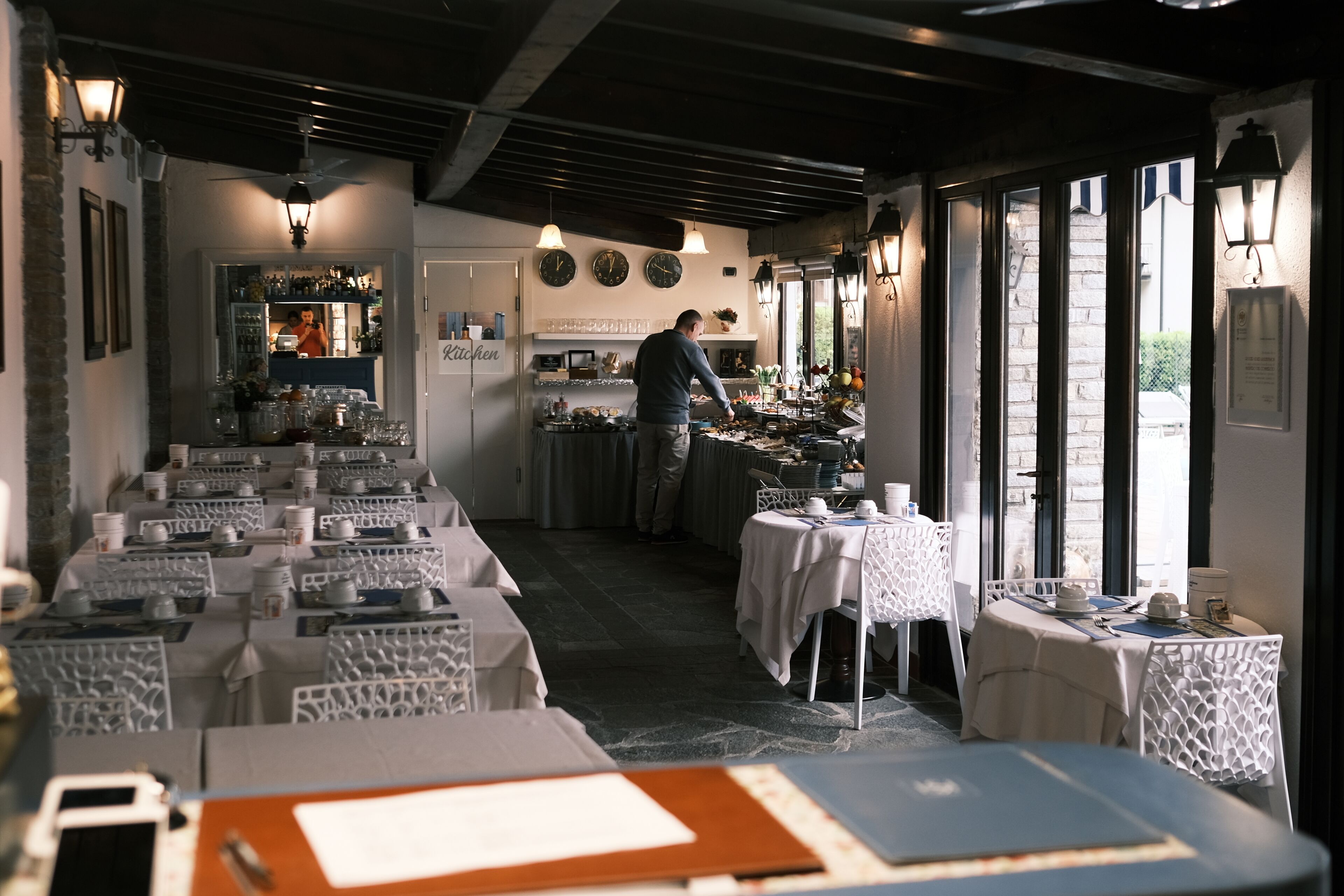 Een restaurant met gedekte tafels, een kok in de weer met eten, en uitzicht op de straat door grote ramen.