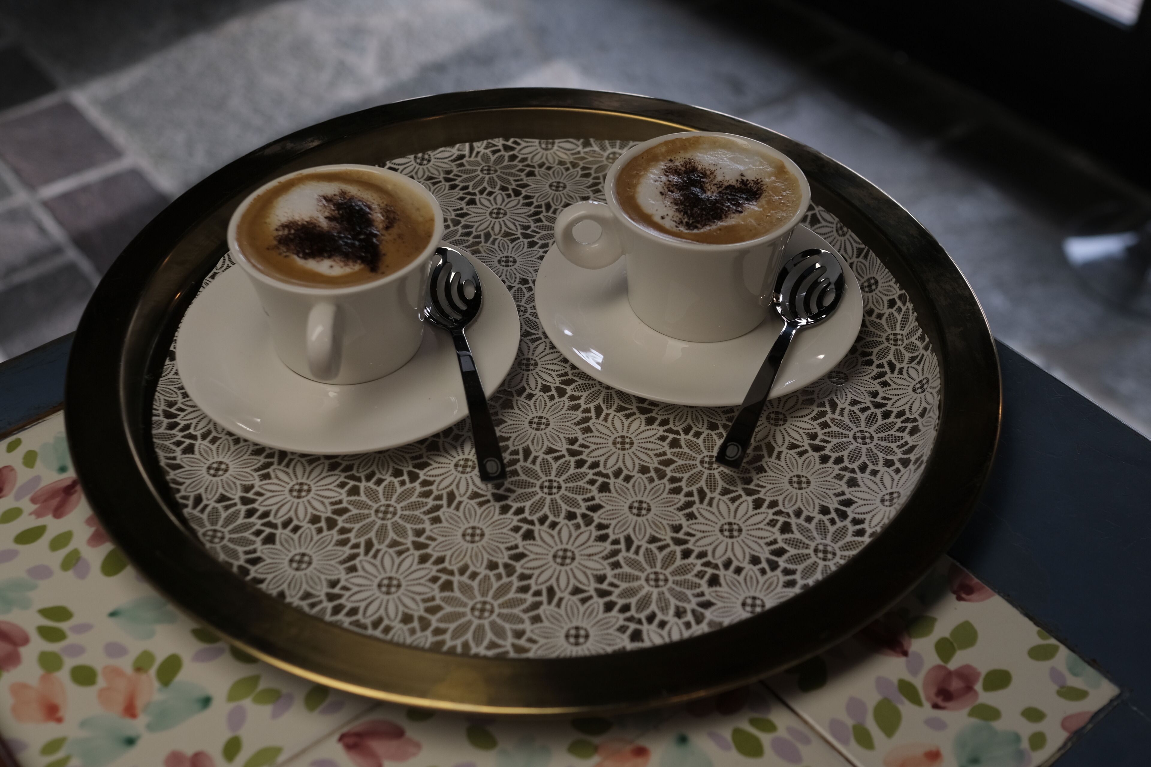 Two cups of coffee on a tray with spoons, placed on a table with a floral tablecloth.