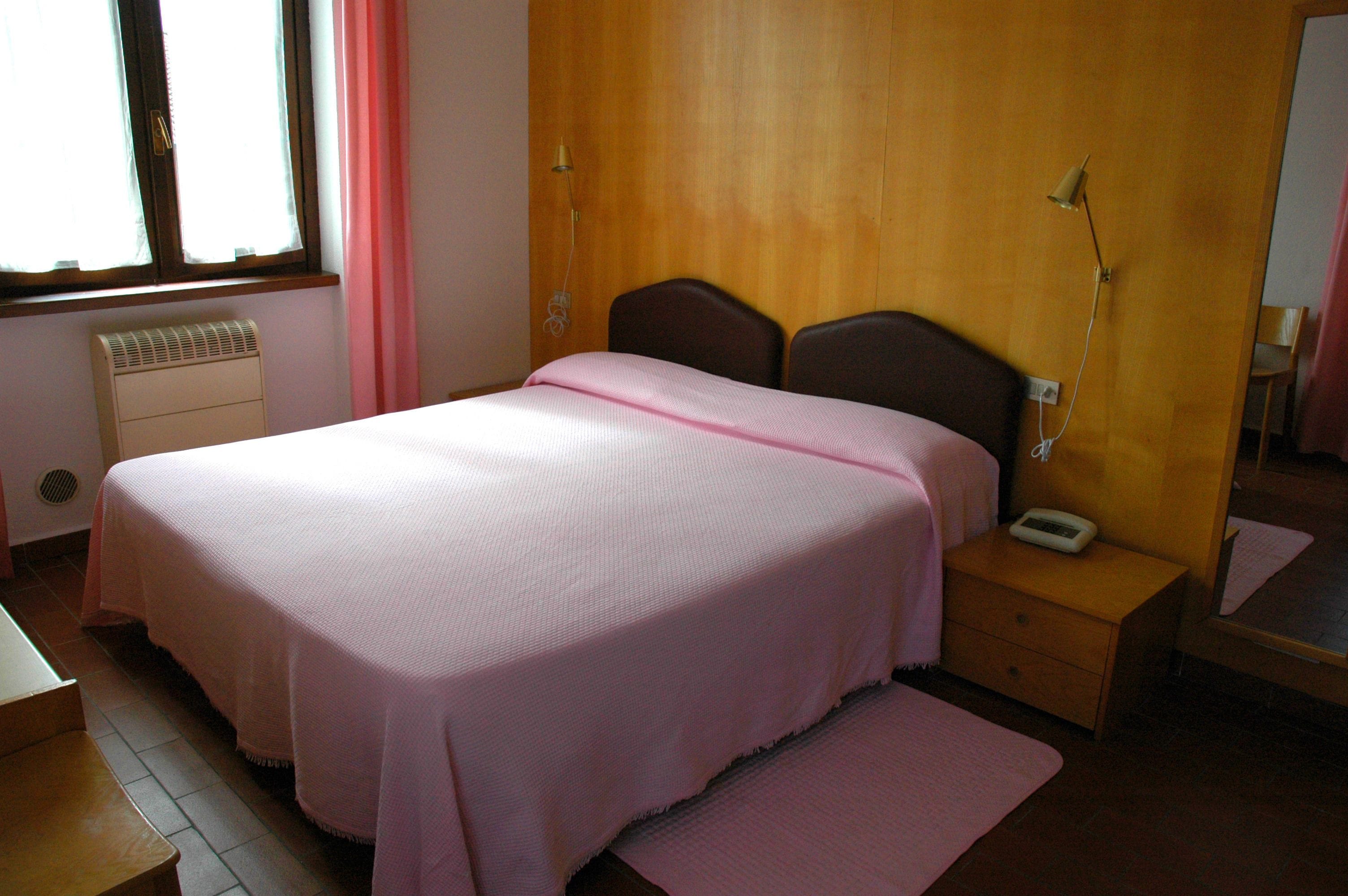 Un dormitorio con cama, mesita de noche, lámpara, calefactor y ventana con cortinas.