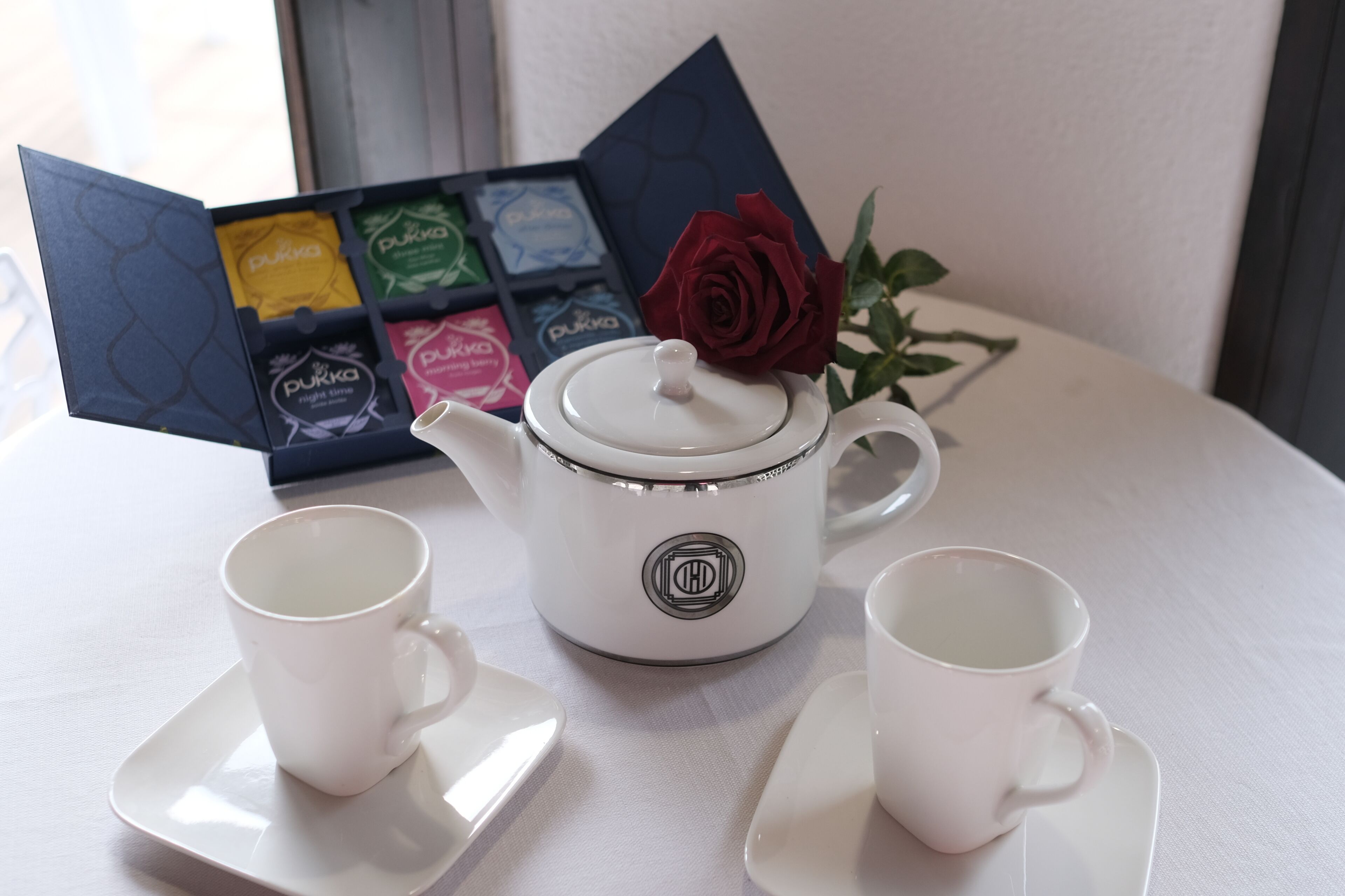 A white teapot with a lid, two white cups on saucers, and a set of tea boxes with a rose on the table.