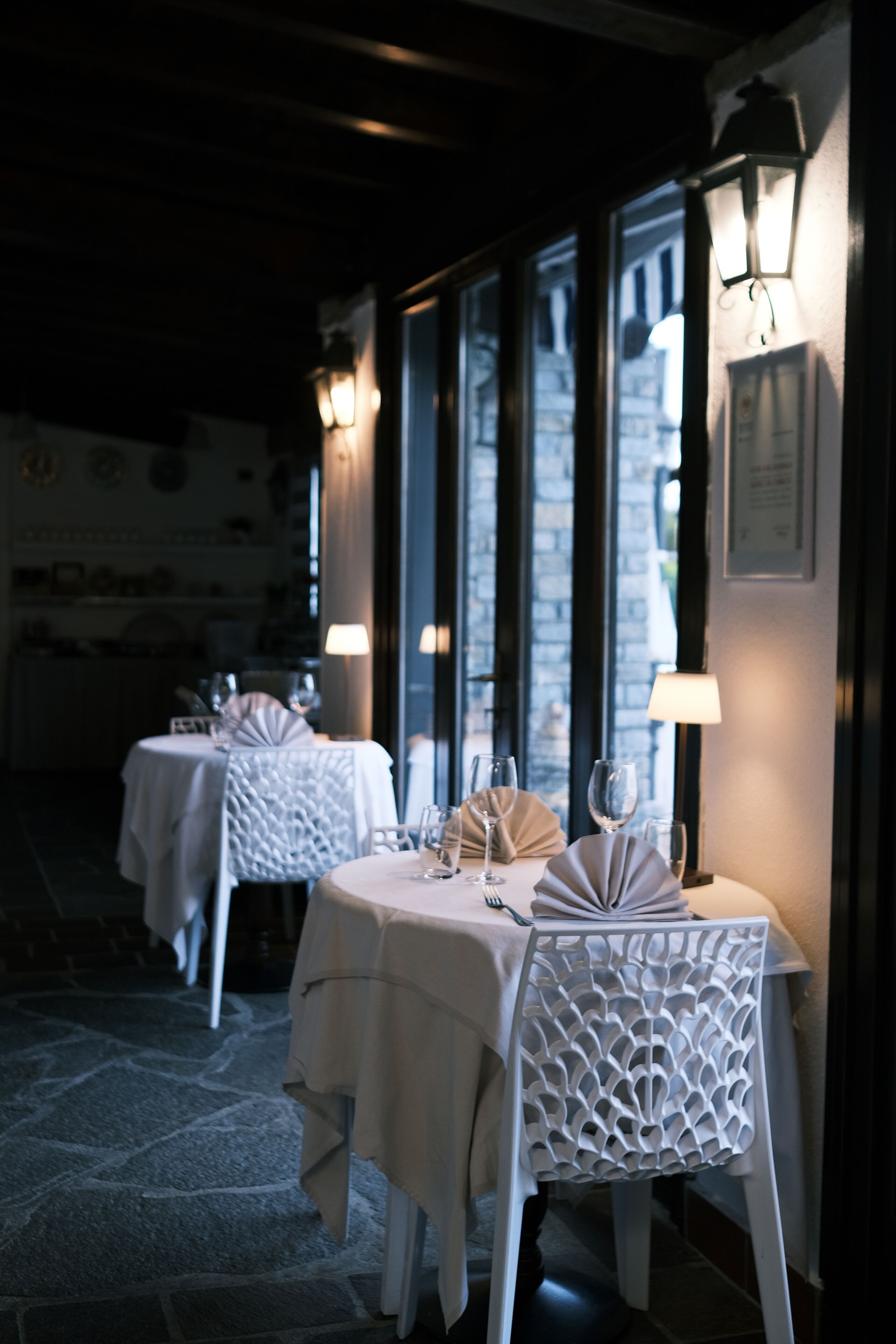 A dimly lit restaurant with tables set for dining, featuring patterned chairs and wall-mounted lamps.