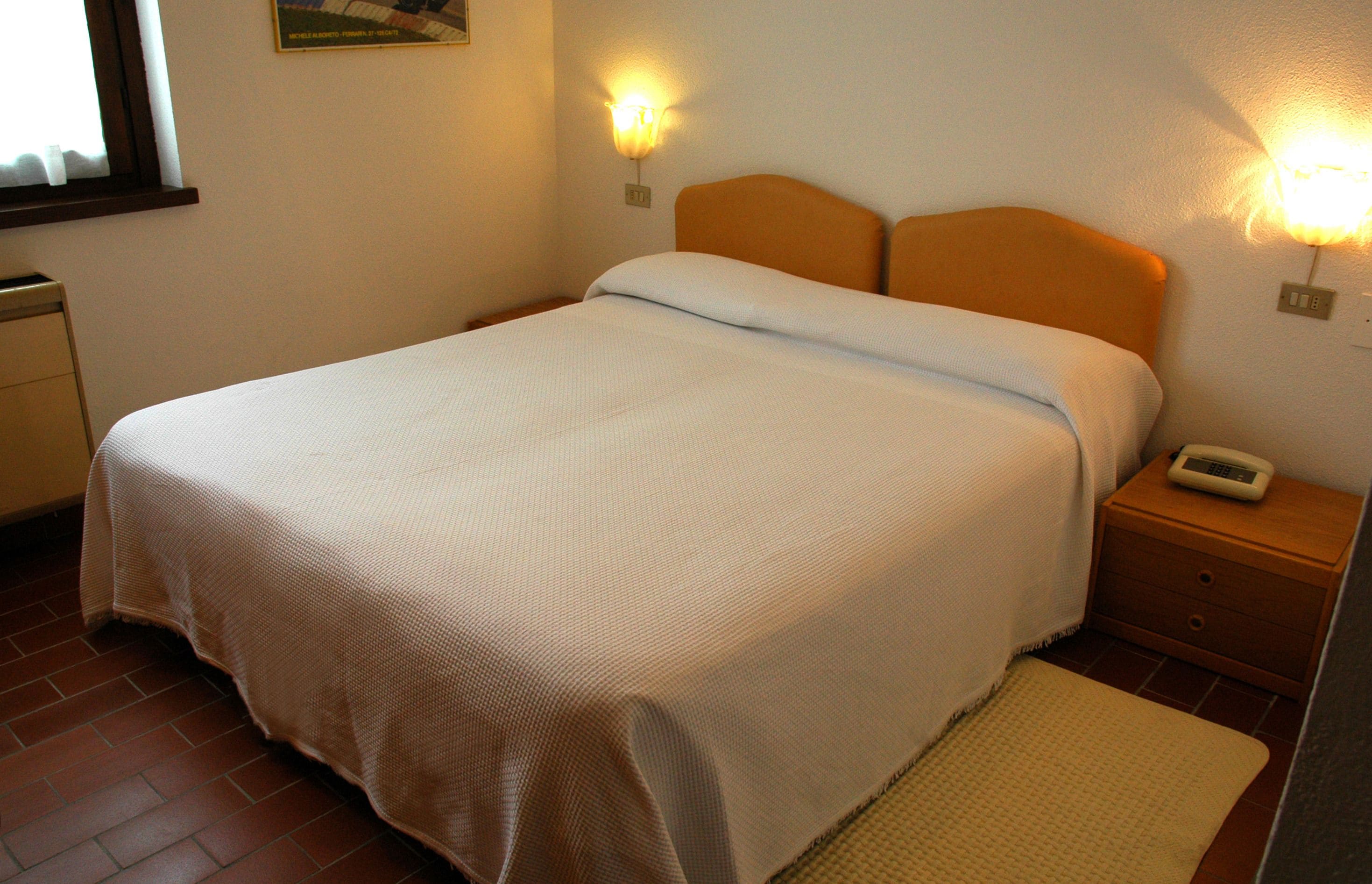 A neatly made bed with a white bedspread, two wall-mounted nightstands with lamps, and a window with curtains.