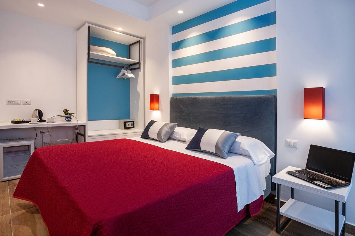 Una habitación de hotel moderna con una cama, un escritorio, una computadora portátil y una alfombra roja.