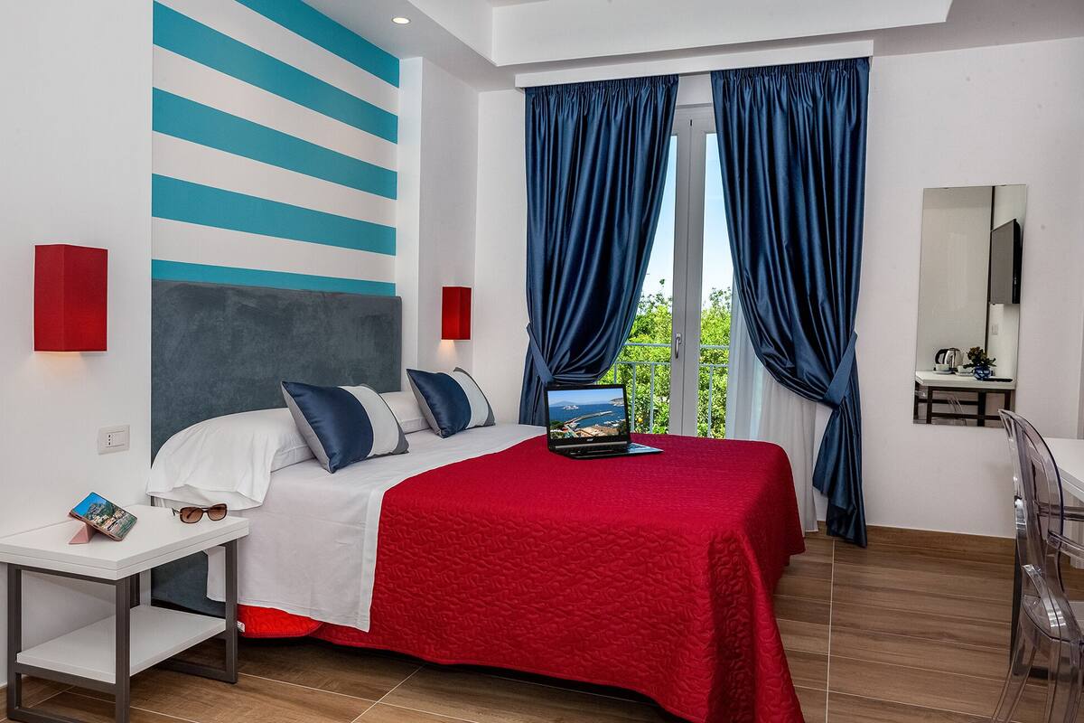Un dormitorio moderno con una cama, una computadora portátil, una colcha roja, una mesita de noche y vistas a la vegetación por la ventana.