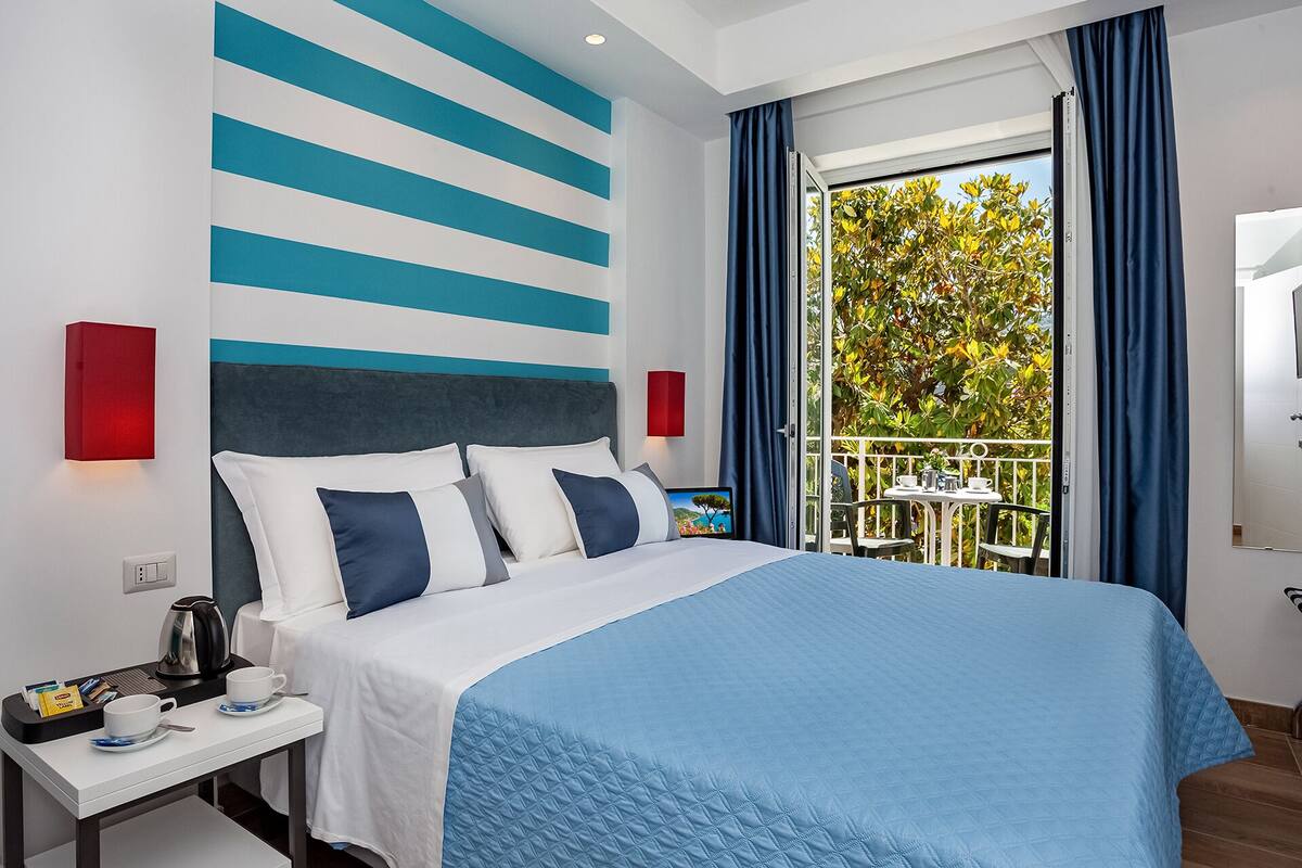 Habitación de hotel moderna con una cama grande, una mesita de noche con cafetera, vistas al exterior y un balcón con mesa y sillas.