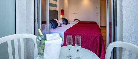 A hotel room with a bed, a red bedspread, a nightstand with a bottle and glasses, and a table with a bottle, glasses, and sunglasses.