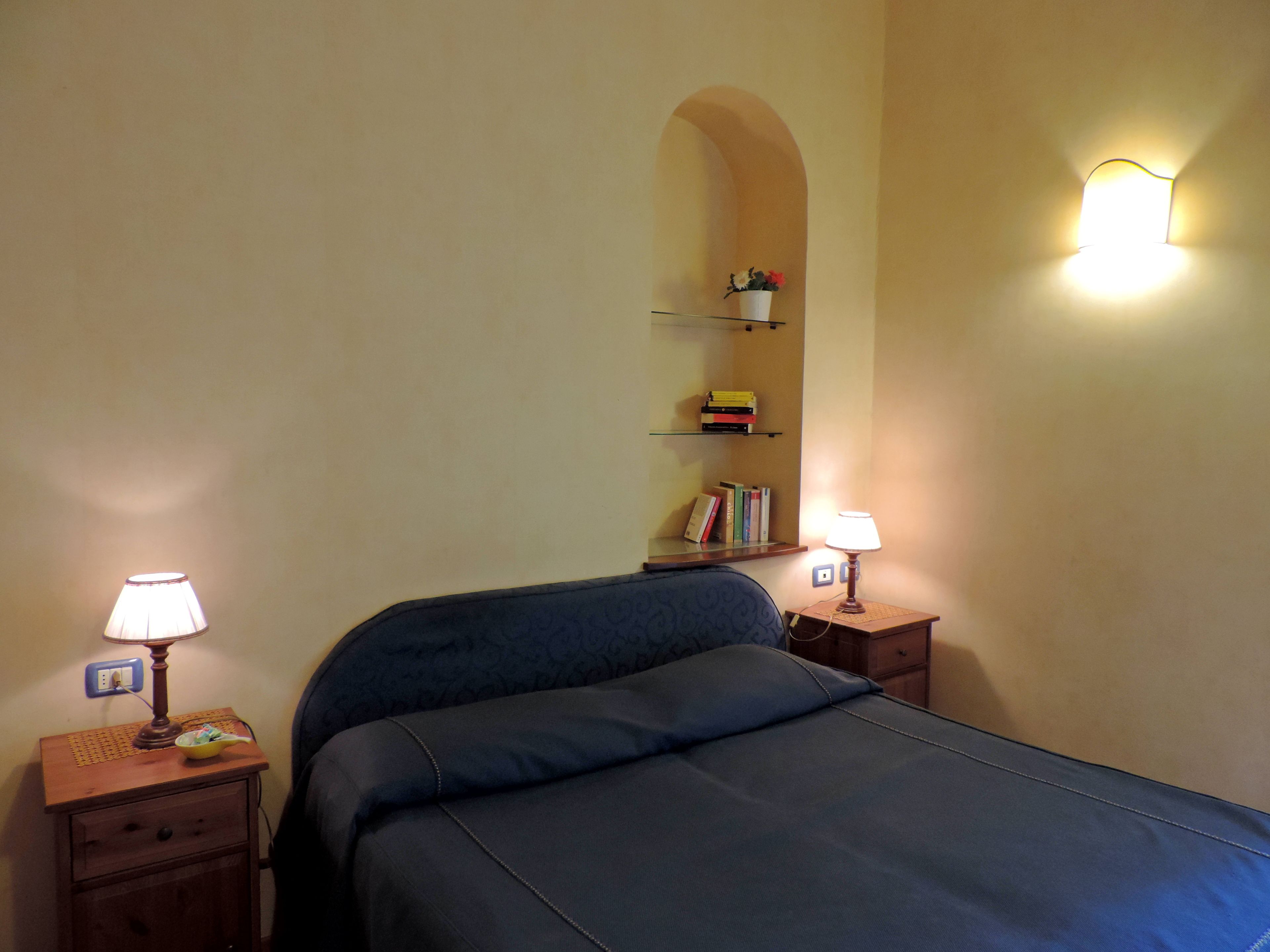 A bedroom with a bed, two bedside tables with lamps, a shelf with books, and a wall-mounted light fixture.