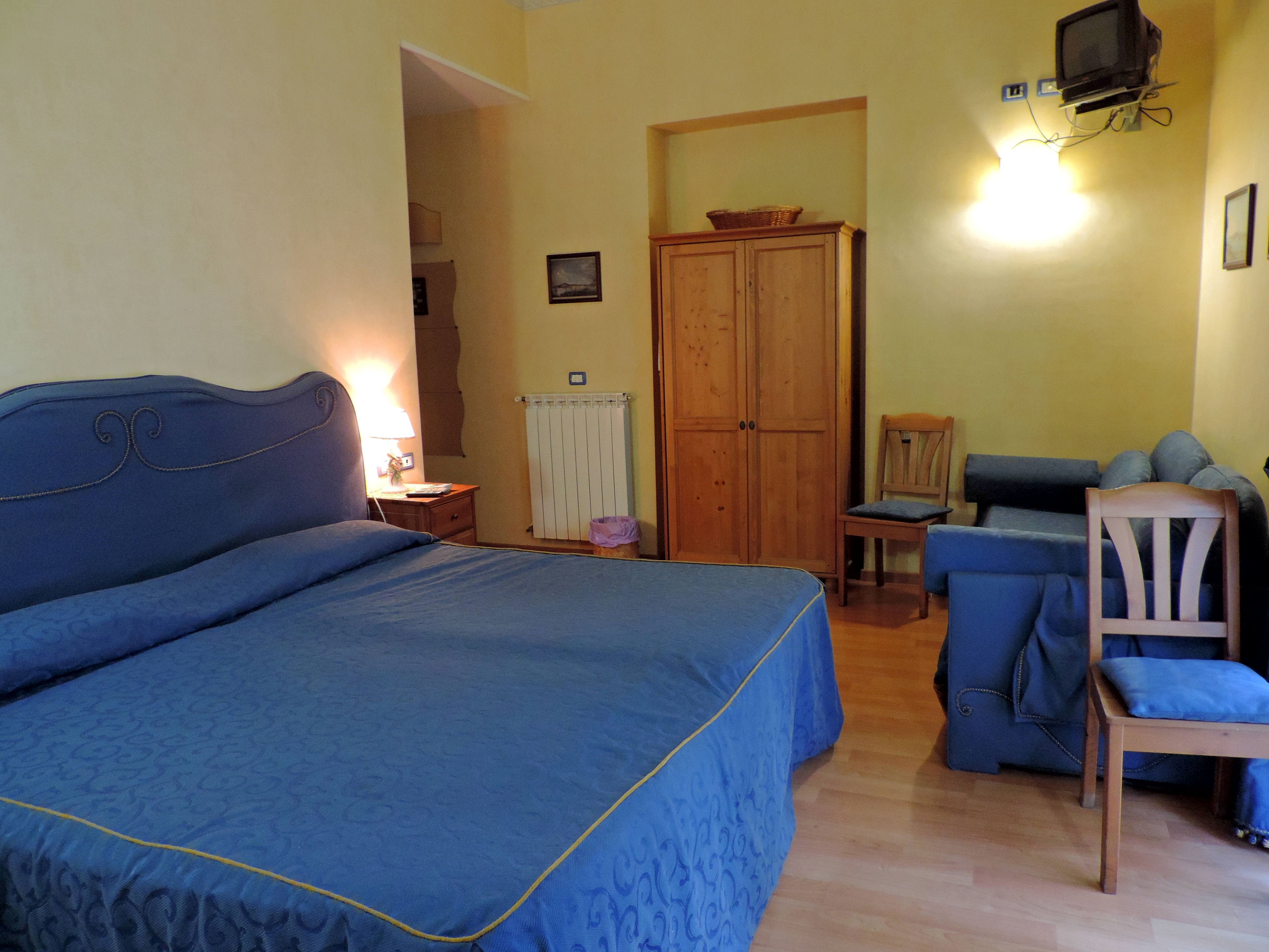 A bedroom with a large bed, a wooden wardrobe, a chair, and a television mounted on the wall.