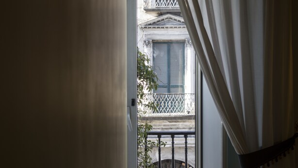 Vista a través de una ventana con cortinas blancas, que muestra un balcón con barandilla metálica y un edificio de arquitectura clásica.