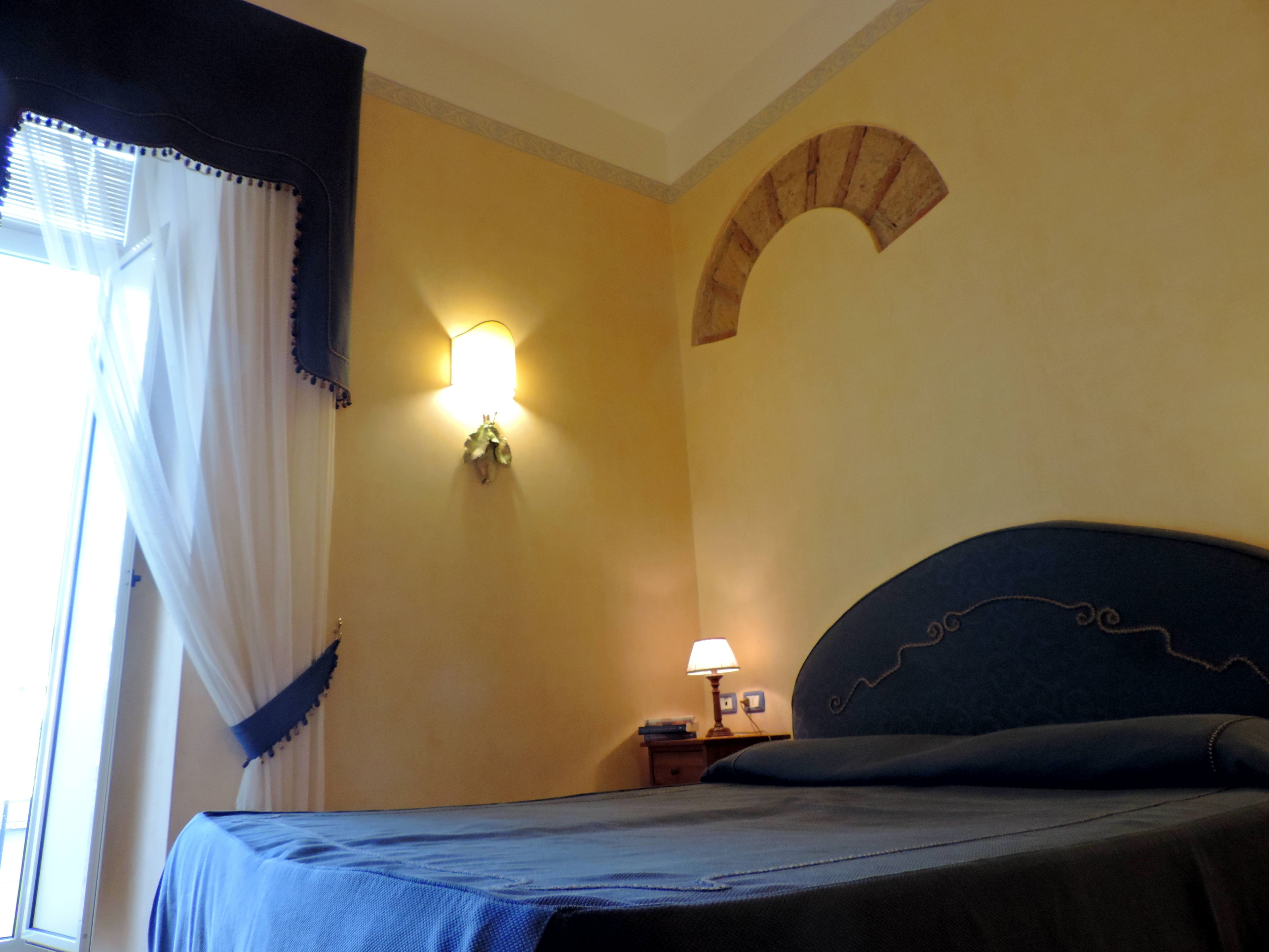 A bedroom with a bed, a nightstand, a lamp, a window with curtains, and a wall-mounted light fixture.