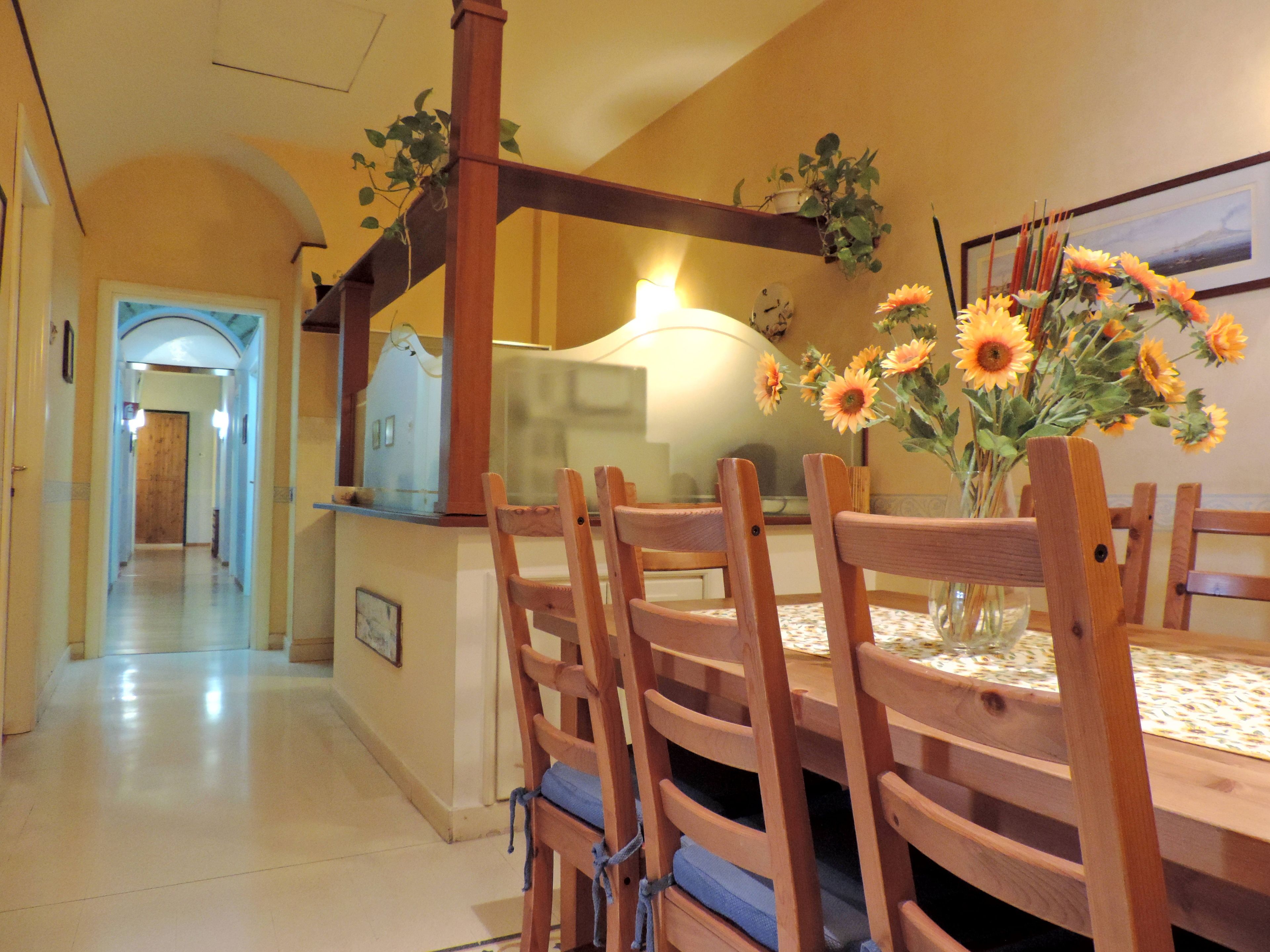 A dining area with wooden chairs and a table, a vase with sunflowers, and a staircase in the background.