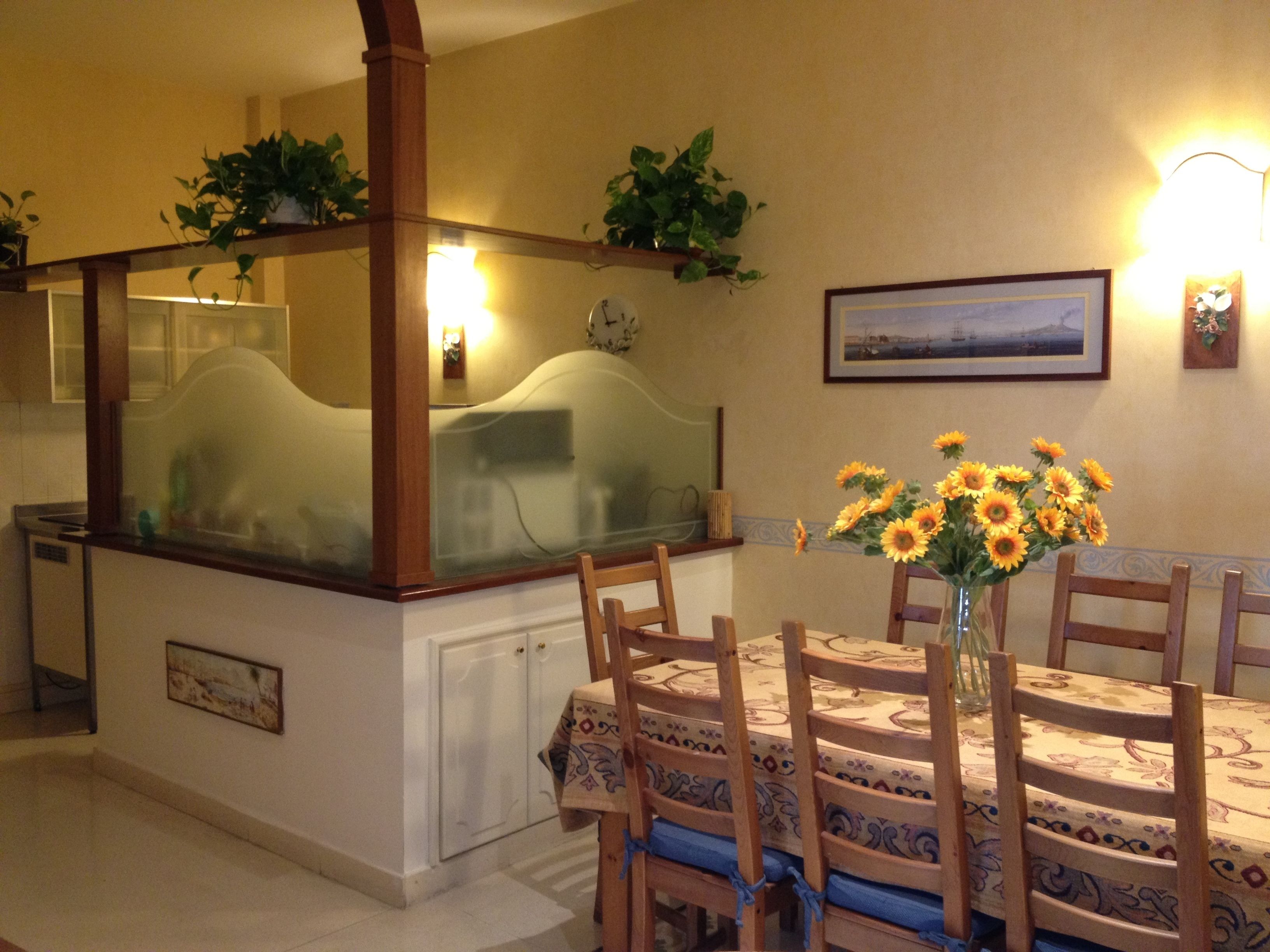 Un espace repas avec un îlot central, des chaises, une table recouverte d’une nappe à motifs, un vase avec des tournesols, des appliques murales et un tableau encadré accroché au mur.