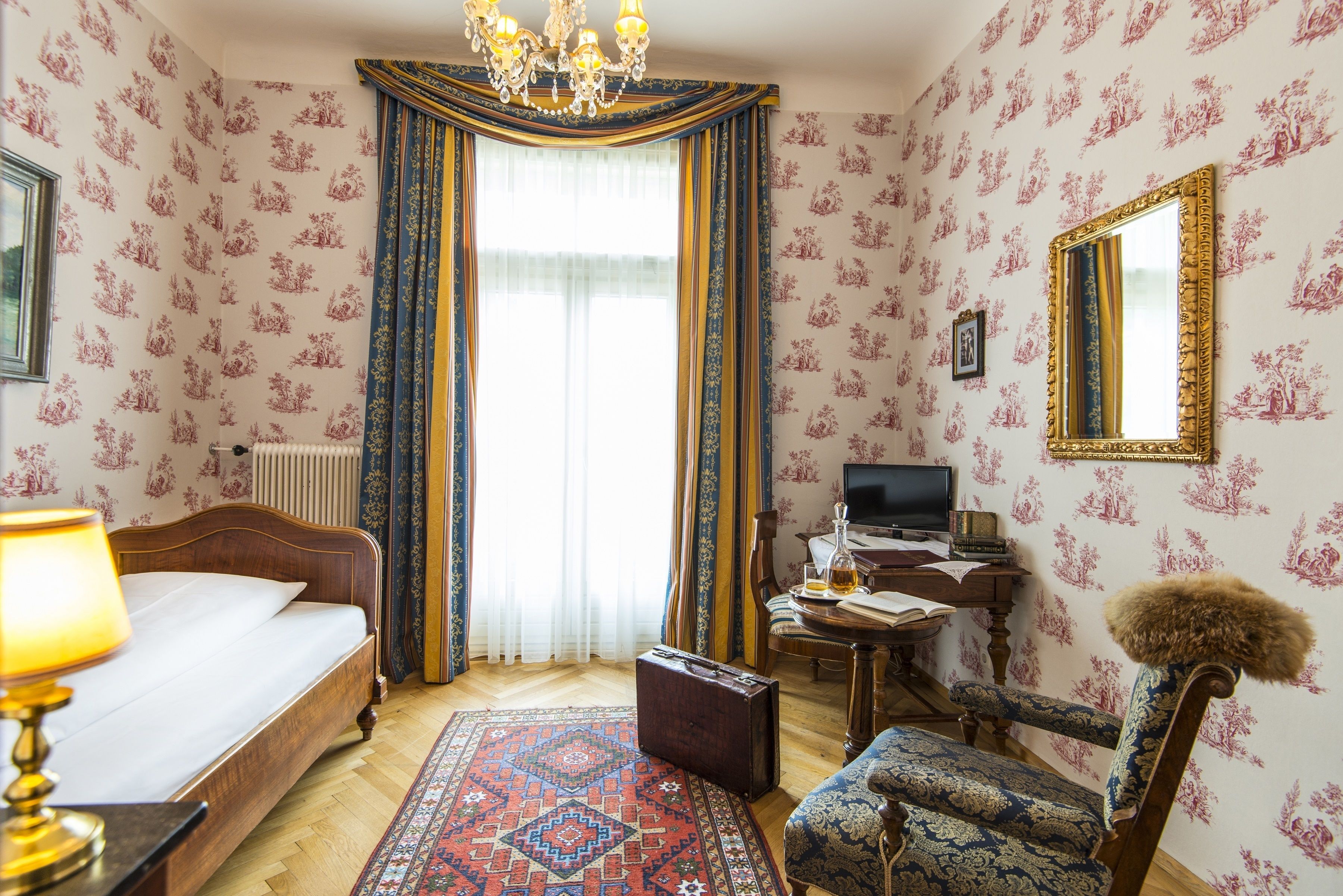 Una camera d'albergo con un letto, una scrivania con un computer, una sedia, un tappeto e un grande specchio.