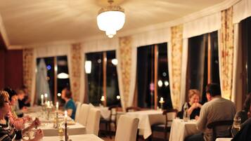 Un restaurante con mesas dispuestas para comer, ventanas amplias con cortinas y un candelabro.