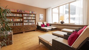 A cozy living room with a large bookshelf, comfortable seating, and a view of a snowy landscape through a large window. - Villa Excelsior Hotel Kurhaus (Bad Gastein)