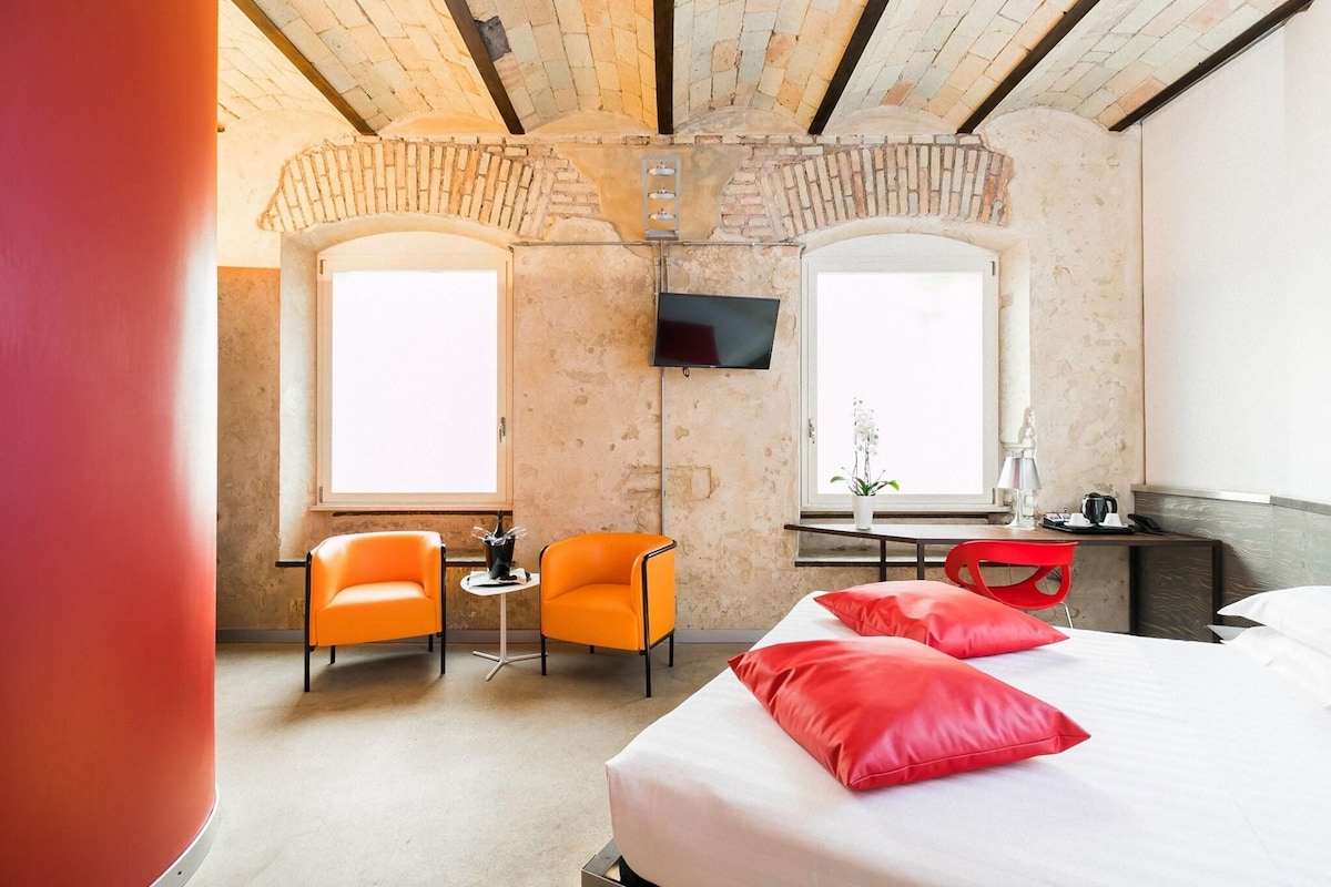 A hotel room with a bed, two orange chairs, a small table, a TV mounted on the wall, and a window with a plant.