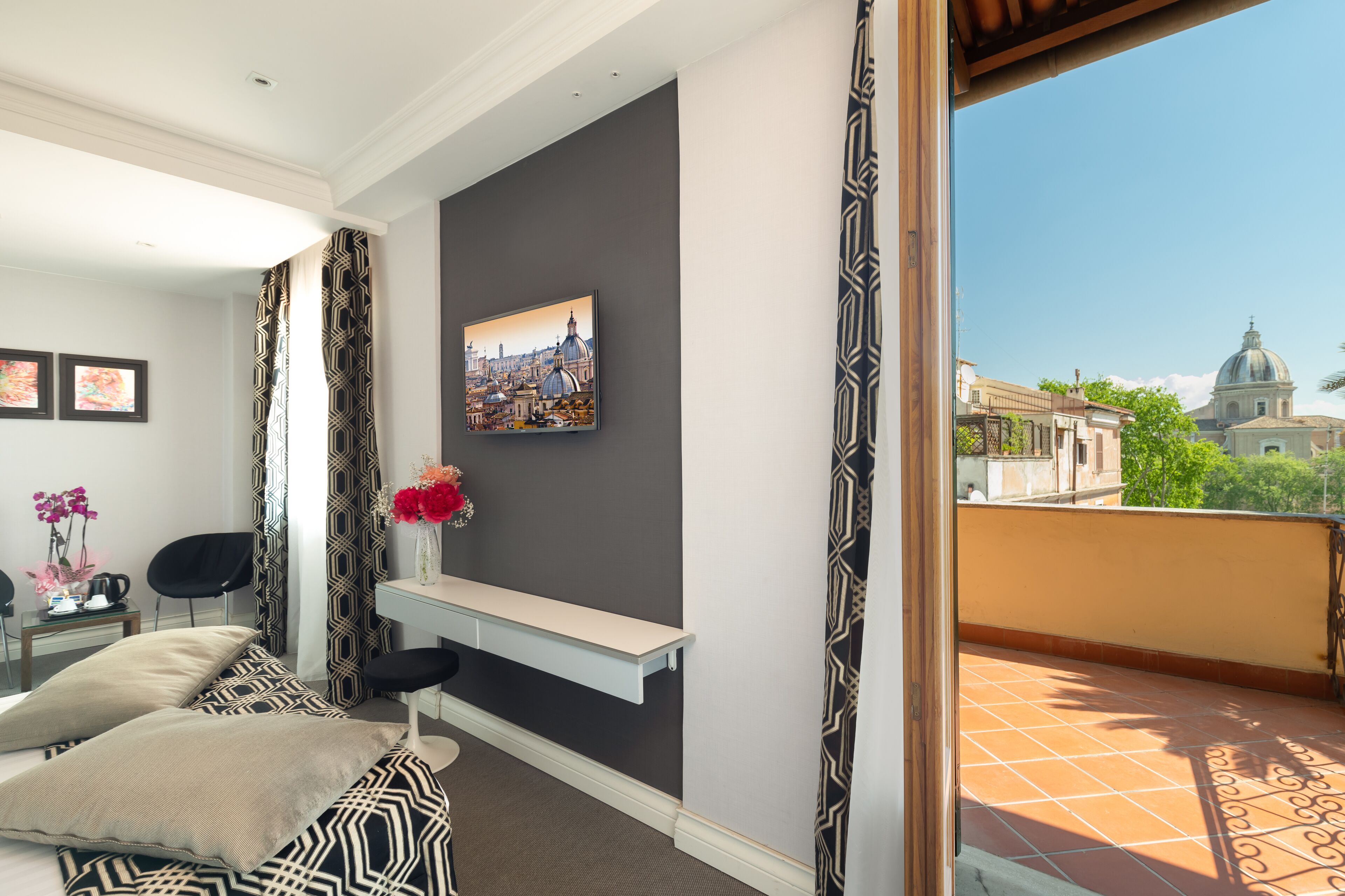 A hotel room with a balcony, a cityscape view, a framed picture on the wall, and a small table with a vase of flowers.