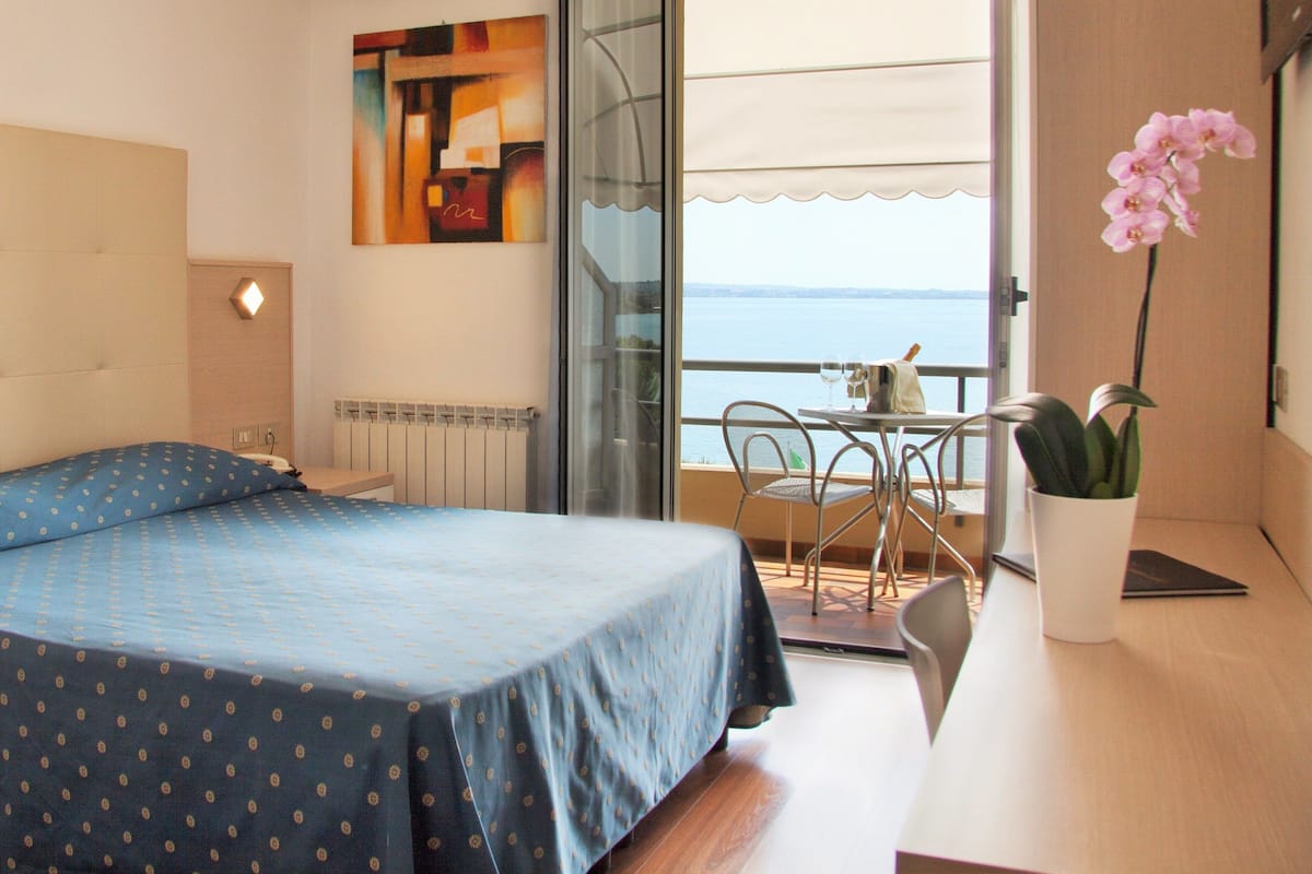 A bedroom with a bed, a desk, a chair, and a view of the sea.