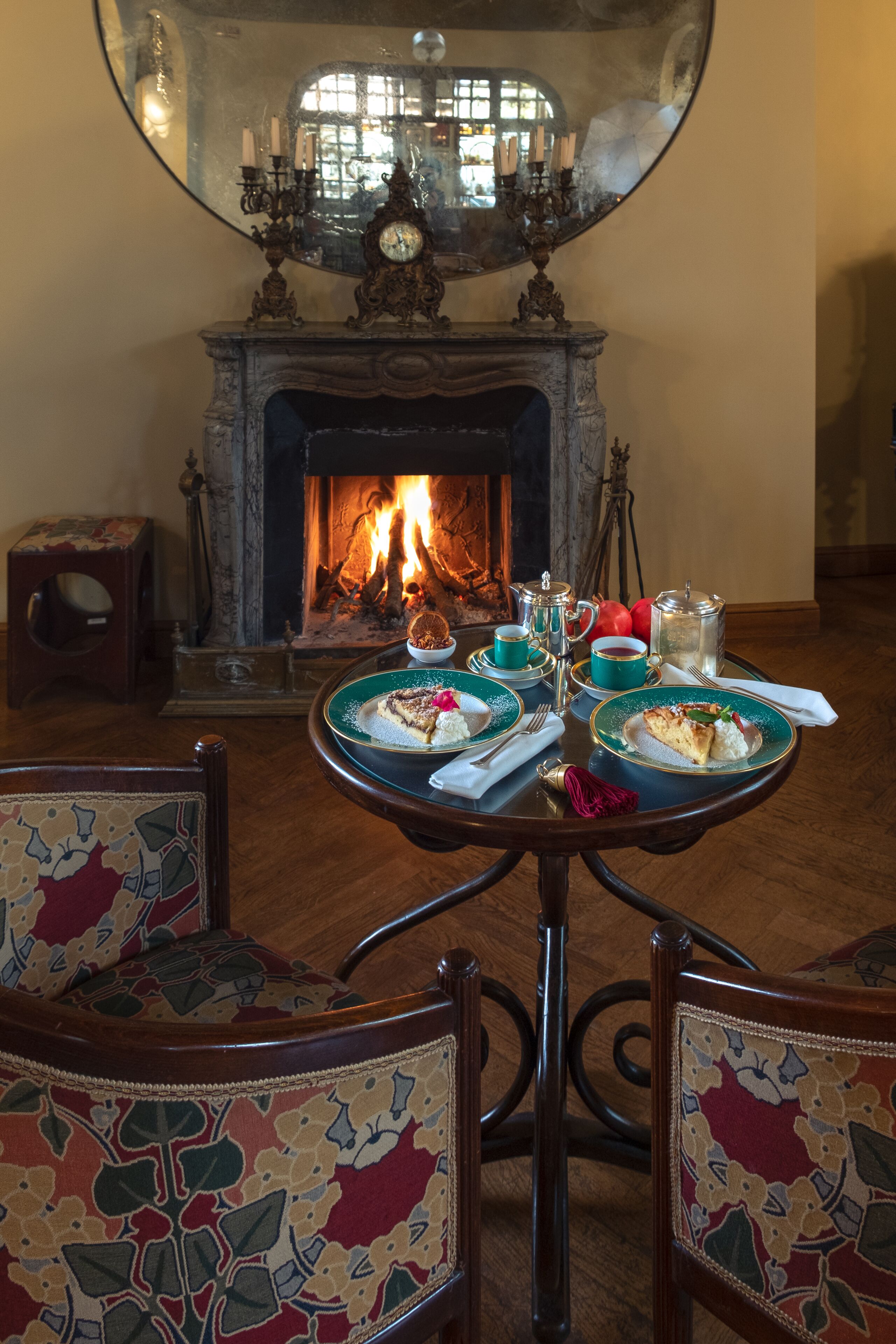 A dining area with a fireplace, ornate mirror, and floral-patterned chairs.