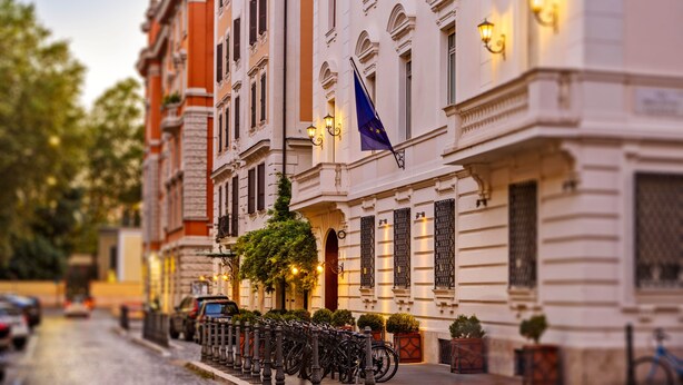 Una fila di edifici storici con una bandiera, un rastrelliera per biciclette e una strada con auto parcheggiate.