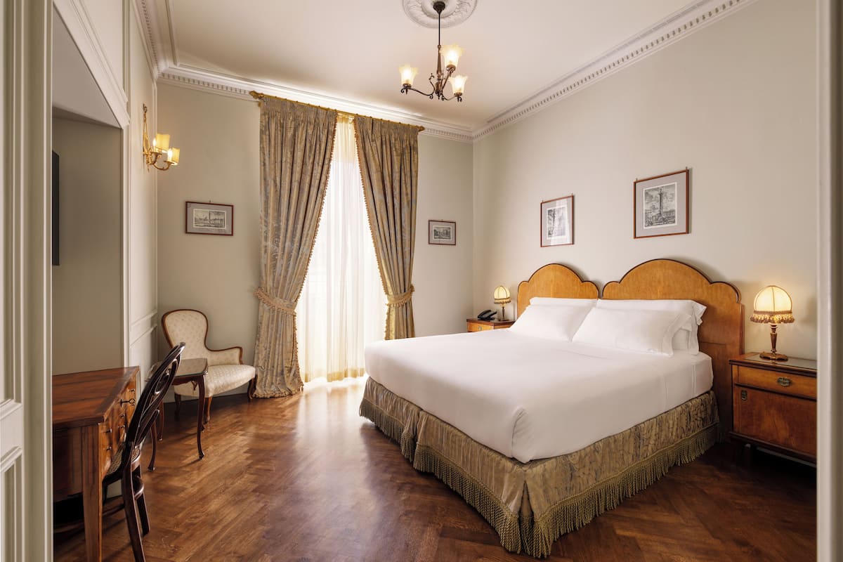 A hotel room with a large bed, wooden headboard, a nightstand with a lamp, a chair, and a desk.