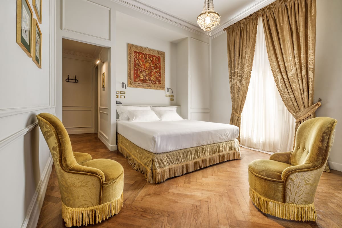 A bedroom with a large bed, two ornate chairs, a chandelier, and a painting on the wall.