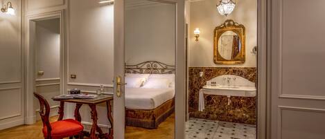 A hotel room with a wooden floor, a bed with white linens, a red upholstered chair, a small wooden table, and a bathroom with a marble sink and mirror.