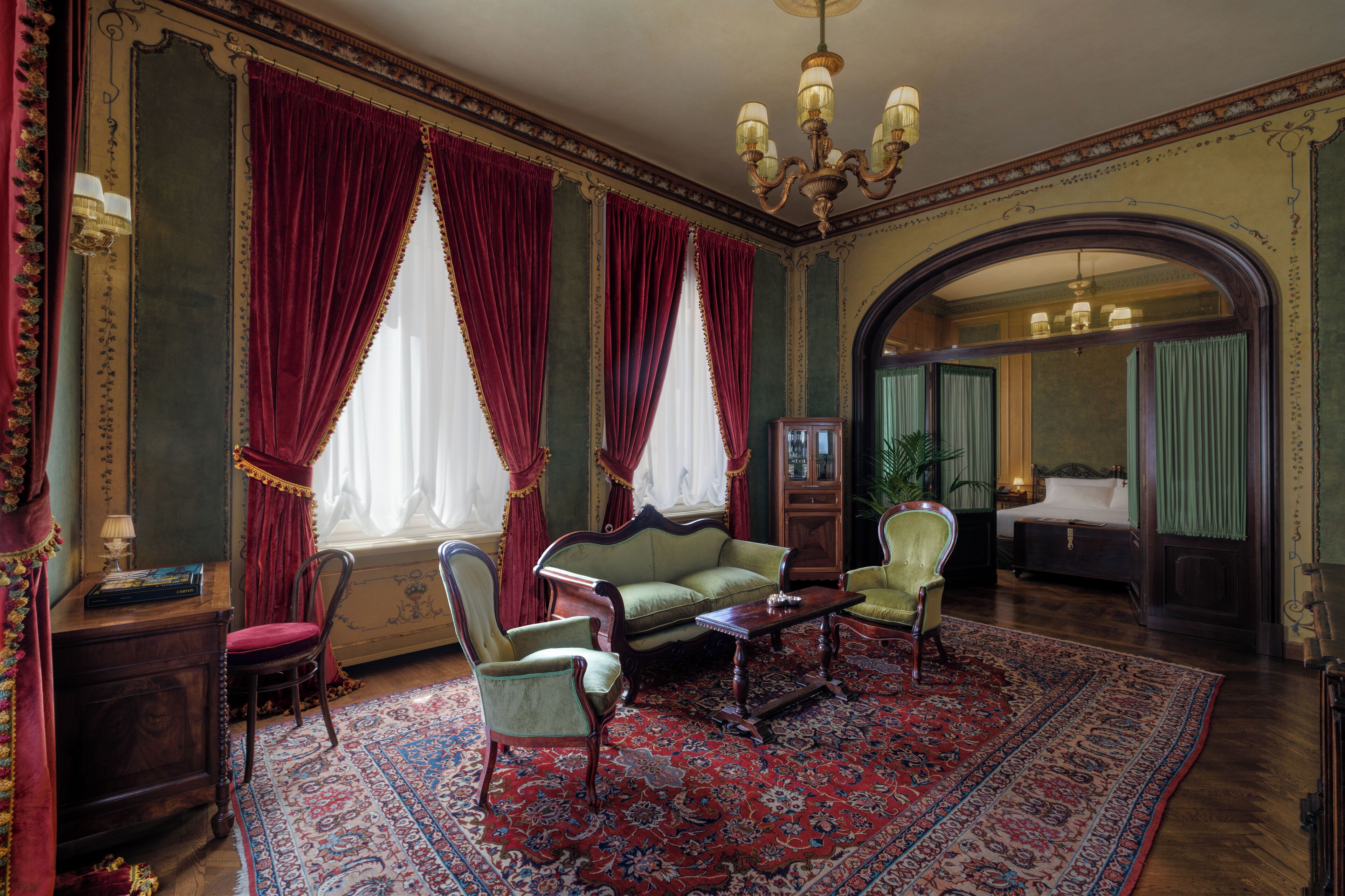 A room with a green sofa, two armchairs, a wooden coffee table, and a red patterned rug. There are two windows with red curtains, a chandelier, and a mirror on the wall.