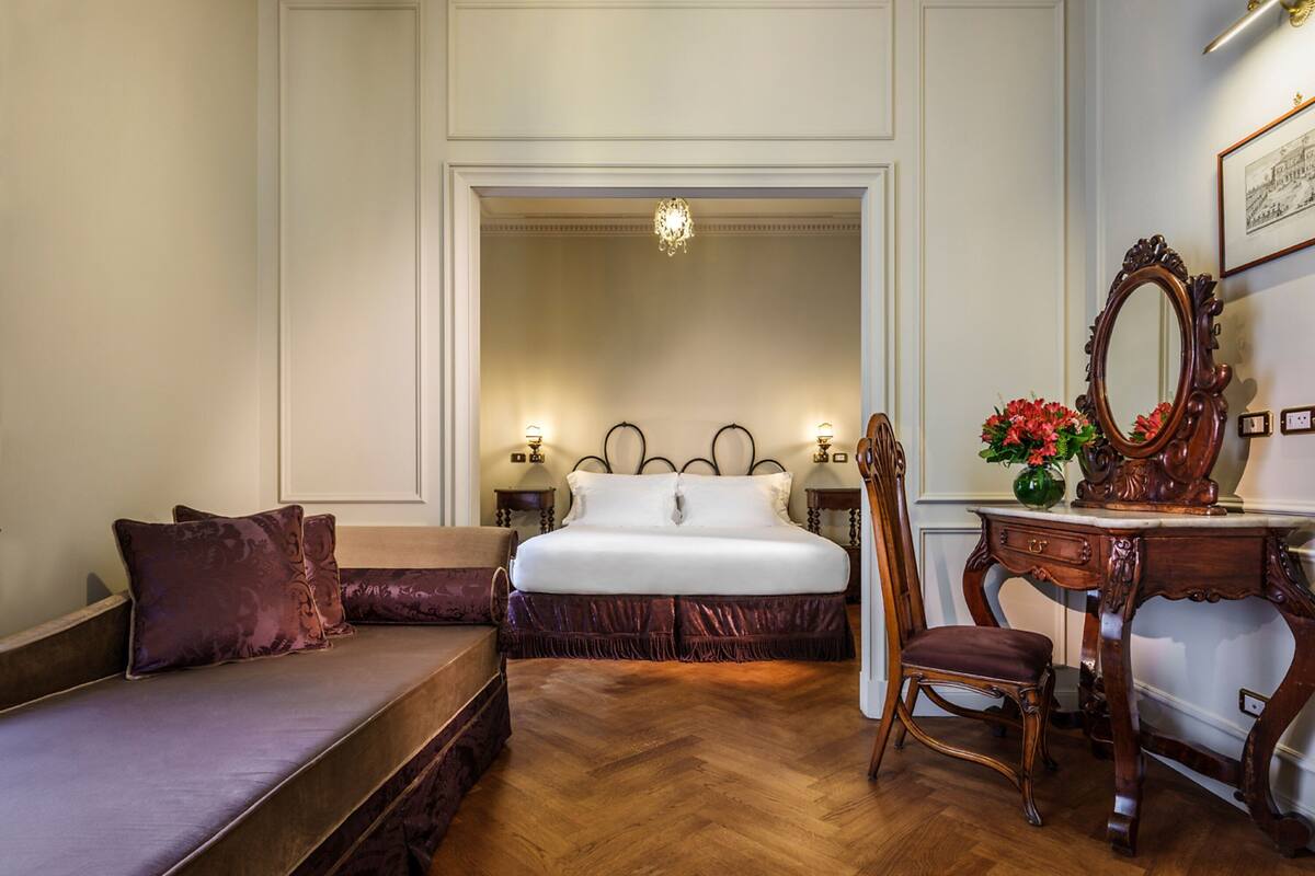 A hotel room with a large bed, a wooden dresser, a vanity with a mirror, and a seating area with a sofa and armchair.