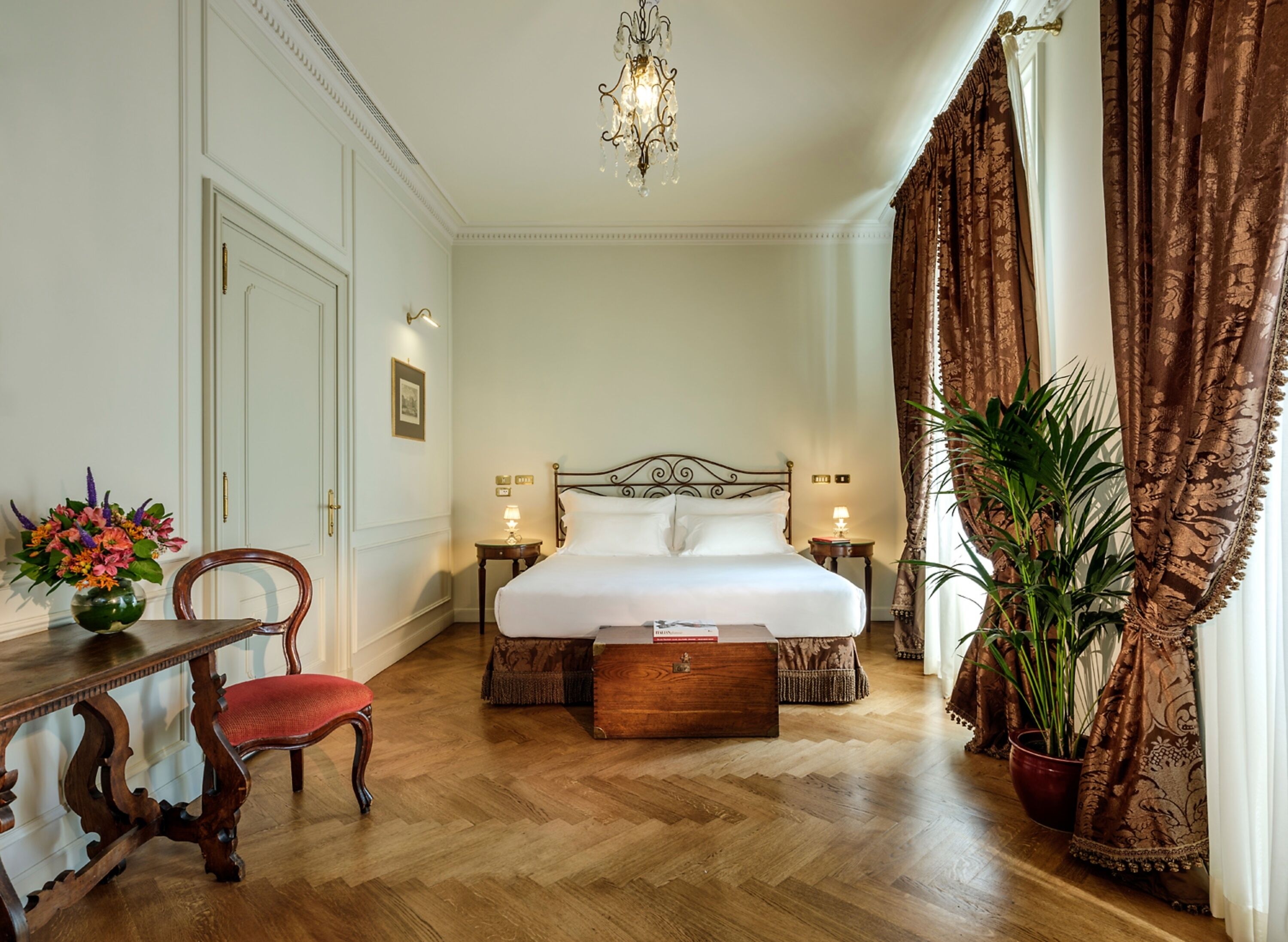 A hotel room with a large bed, a wooden chair, a bedside table, a nightstand, a chandelier, and a potted plant.