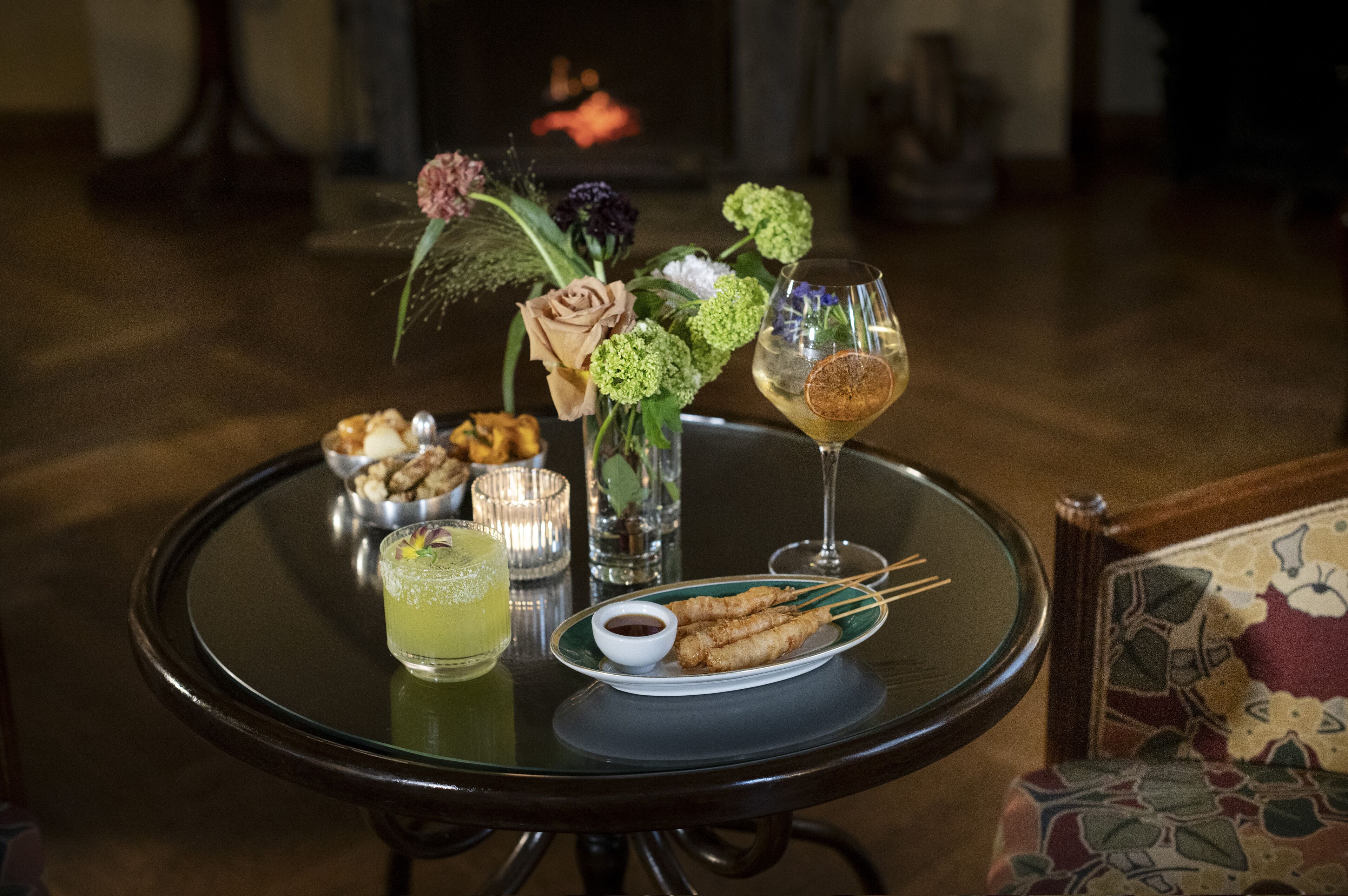 A table with a plate of fried items, a glass of drink, a small bowl of sauce, and a vase of flowers.