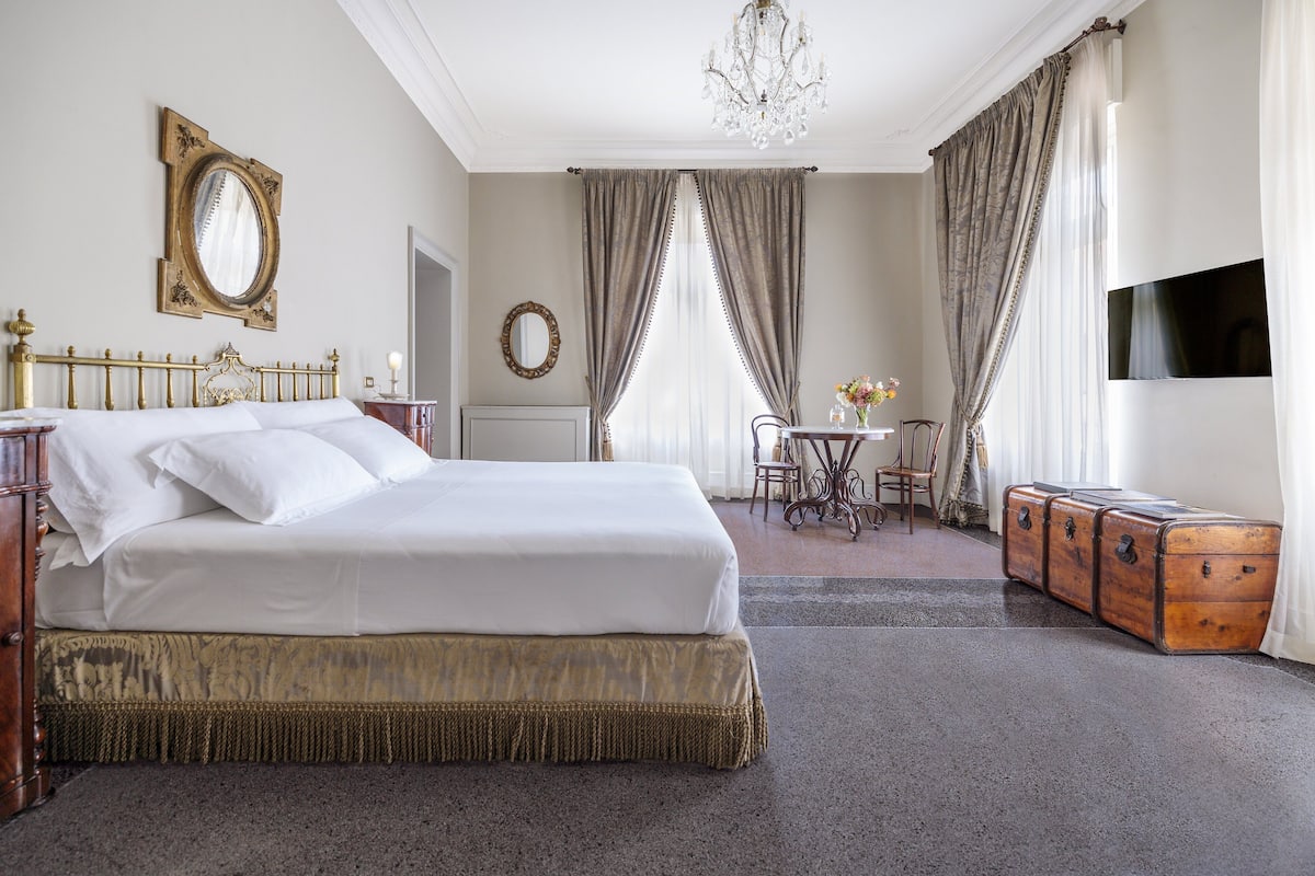 A hotel room with a large bed, a chandelier, a desk with a chair, and a television on the wall.